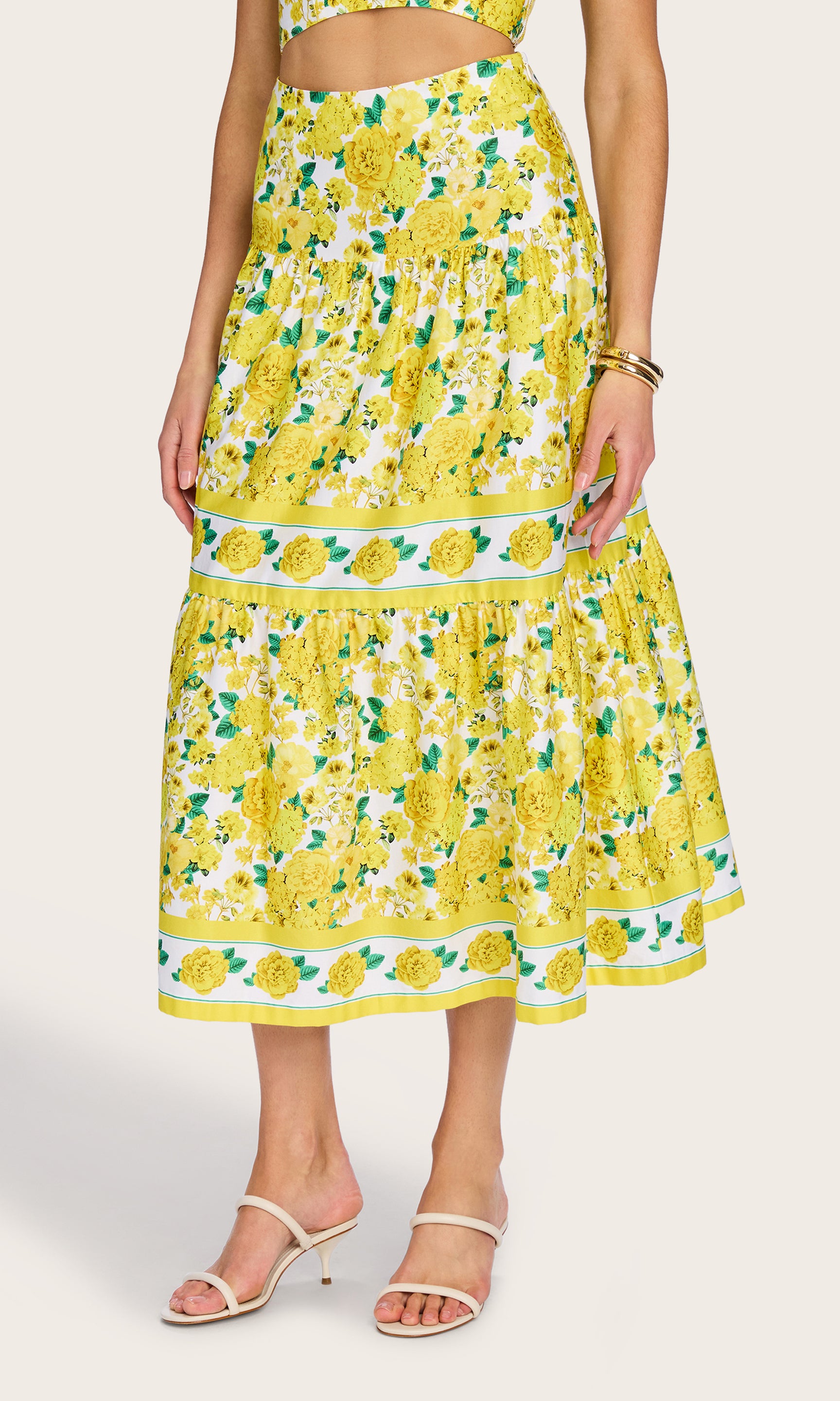 3/4 front view of the model wearing the Mondo Floral Poplin Skirt in a yellow floral print with the matching floral bra top, and white heels.