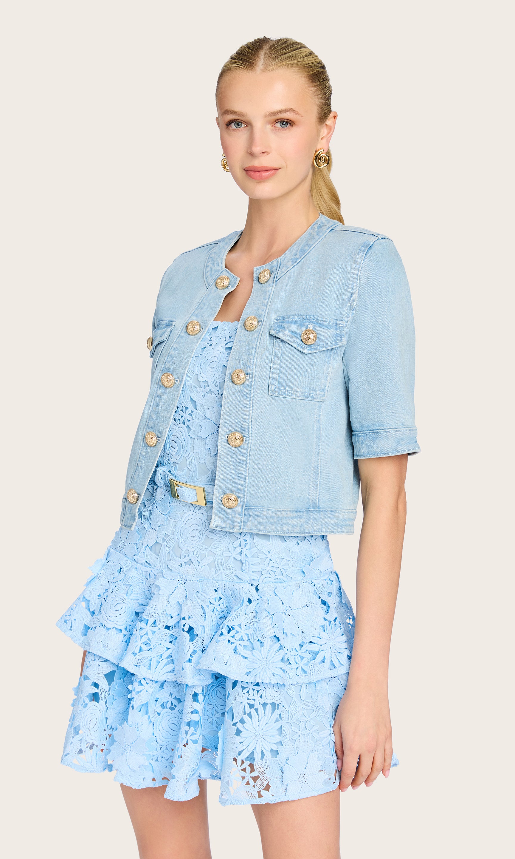 The model wears the Devika Denim Jacket in a clean front view, showcasing its cropped silhouette, decorative buttons, and short sleeves styled with a tiered mini dress.