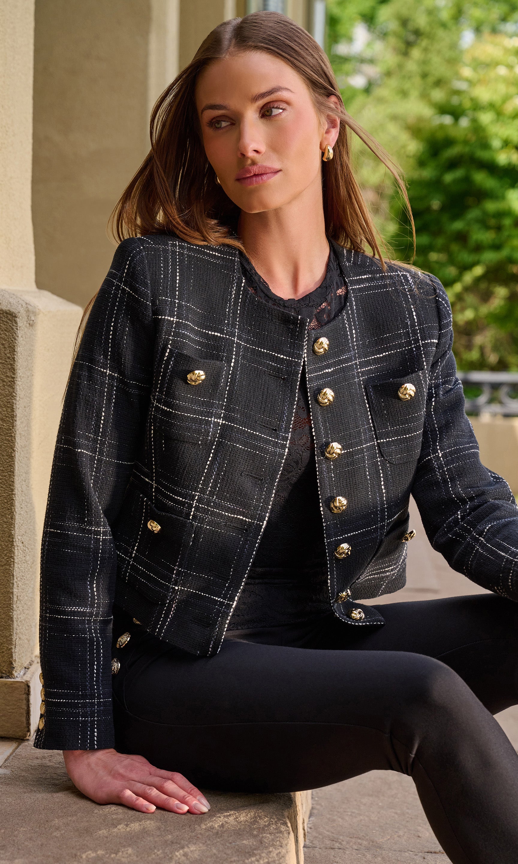 A woman wearing a cropped black plaid jacket with gold buttons, black leggings, and black shoes, sitting on a stone ledge in an outdoor setting.