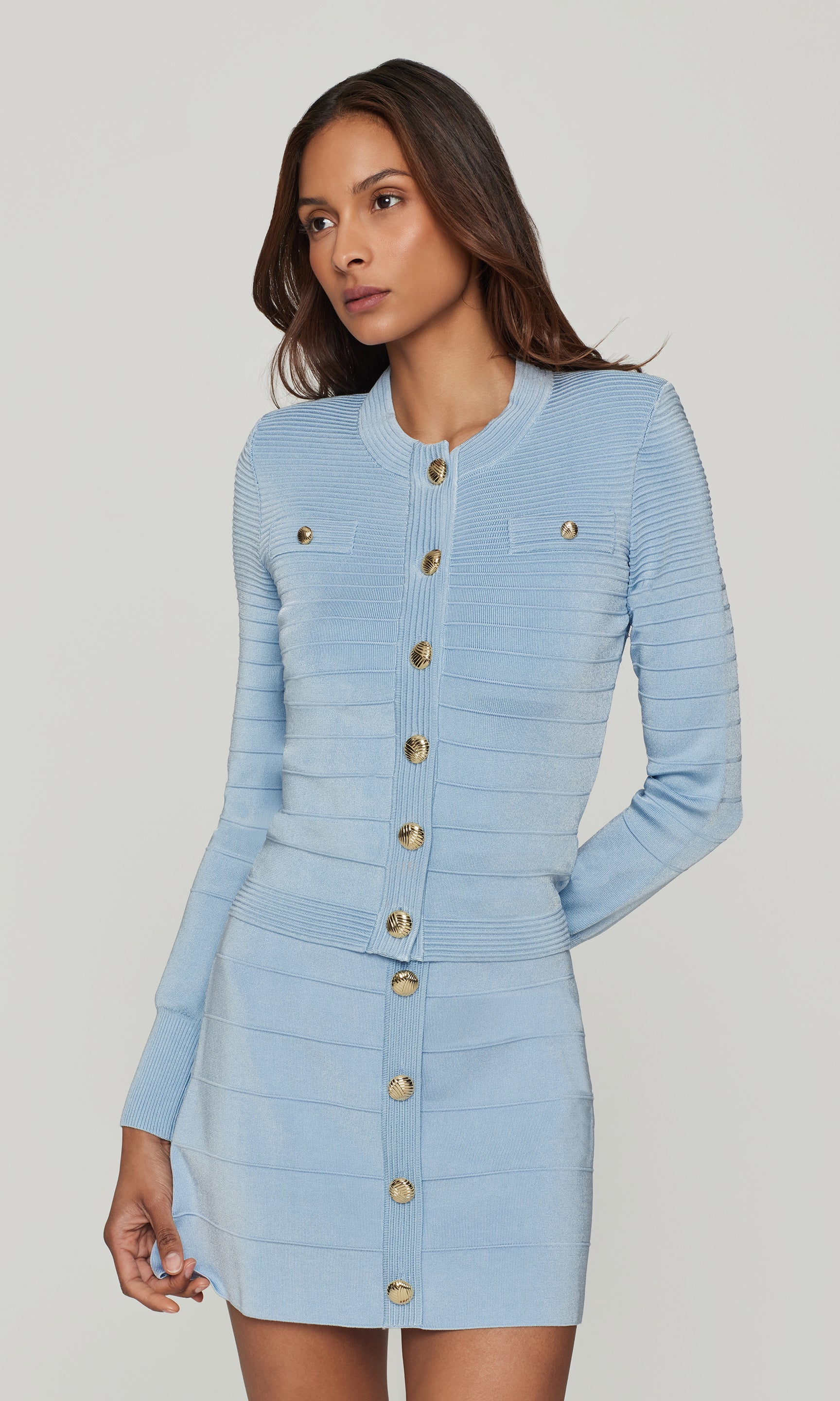 Woman wearing a light blue knitted long sleeve top and matching skirt with gold buttons on a plain background