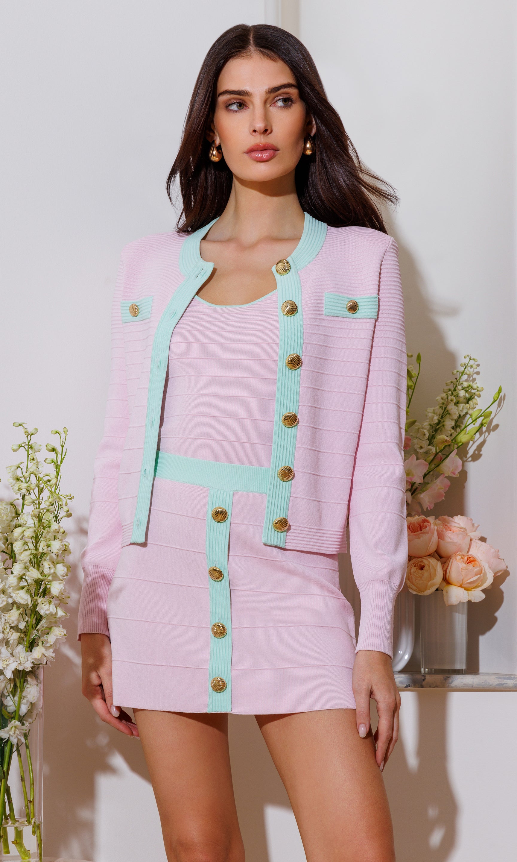 The model wears the Lilla Bandage Cardigan in pink in a three-quarter front view, highlighting the mint trim, gold-tone buttons, and pocket details styled with the matching mini skirt.
