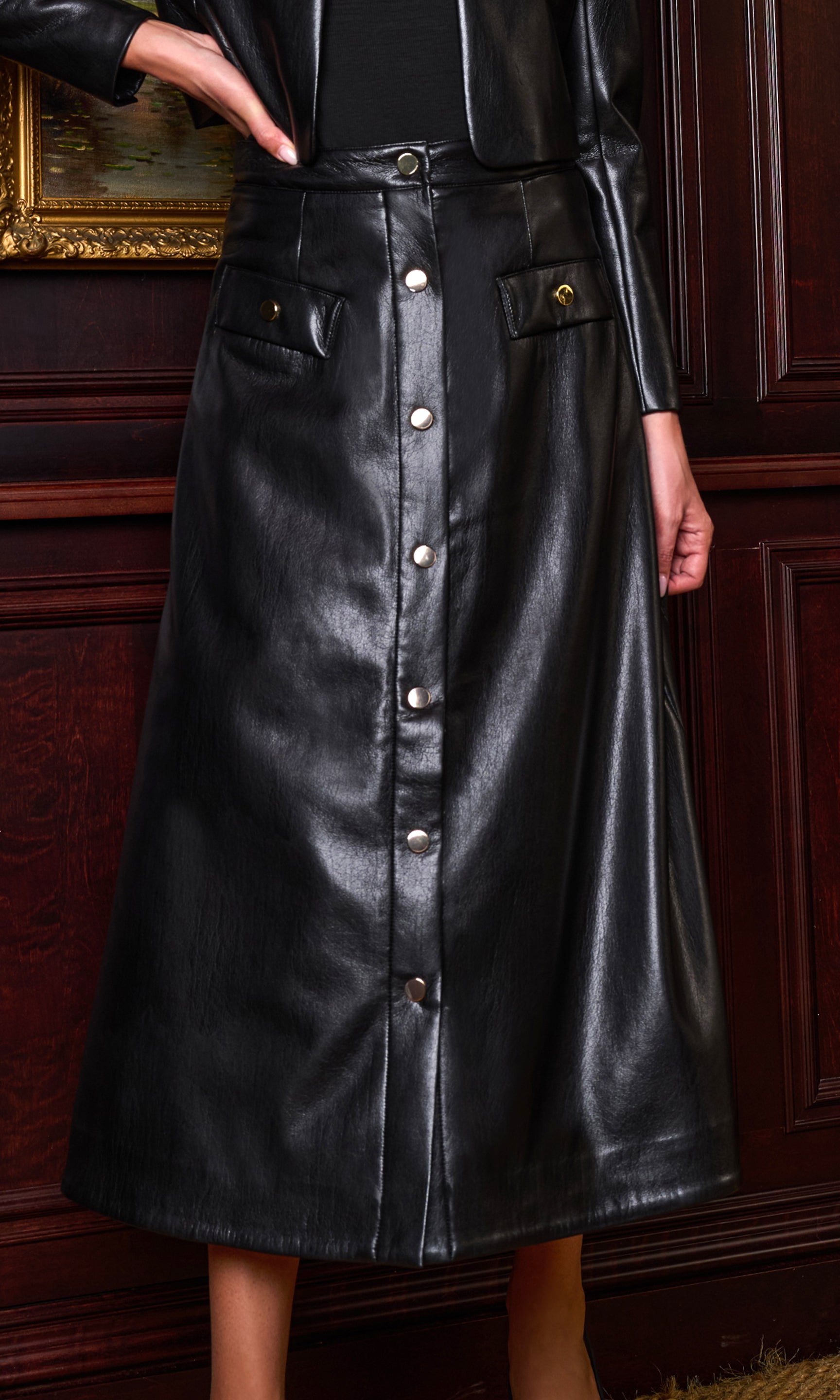 A black faux leather midi skirt with a button-down front and flap pockets, shown on a model in front of a dark wood paneled wall.