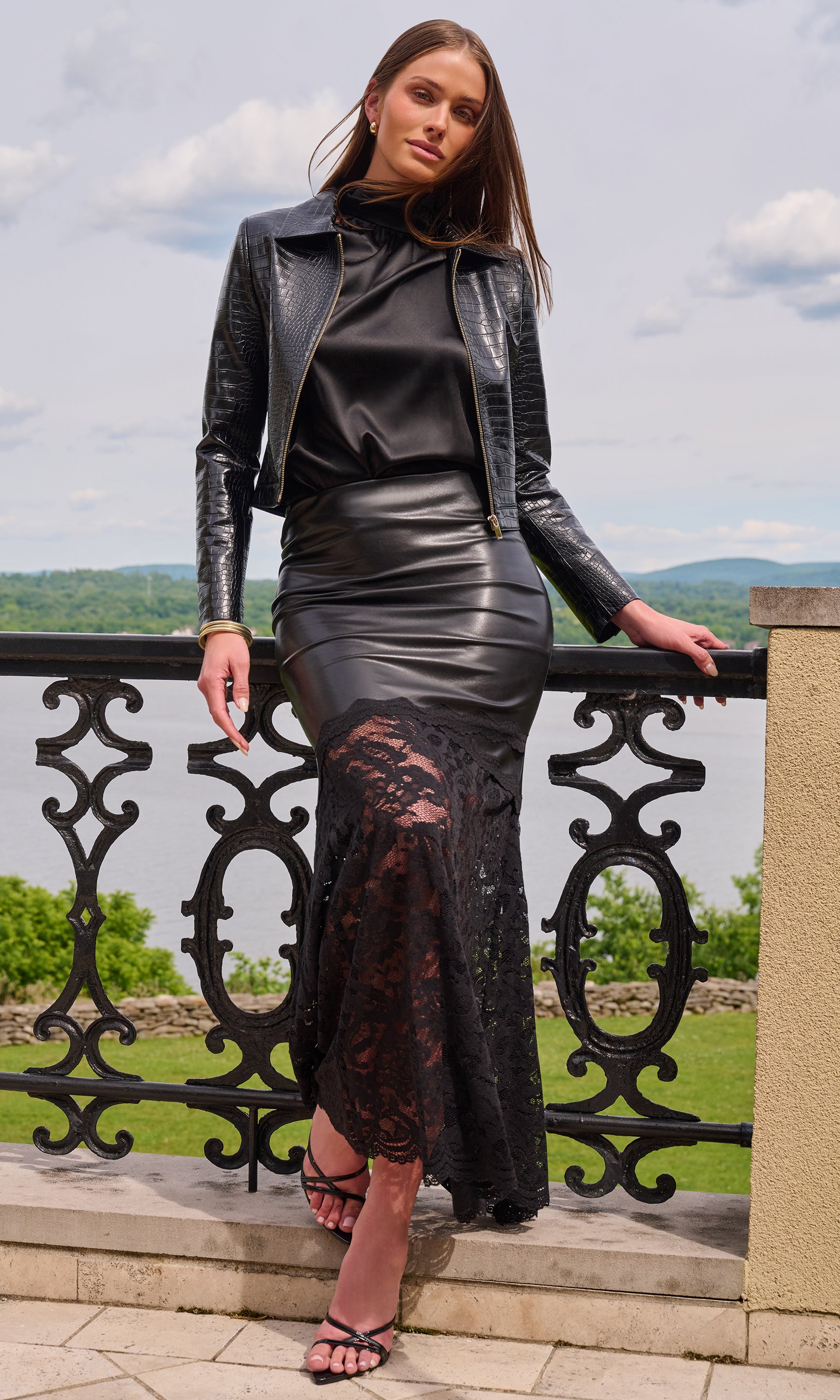 Model stands outdoors on a terrace wearing a black faux-leather moto jacket over a black satin top and a black lace-trimmed pencil skirt.