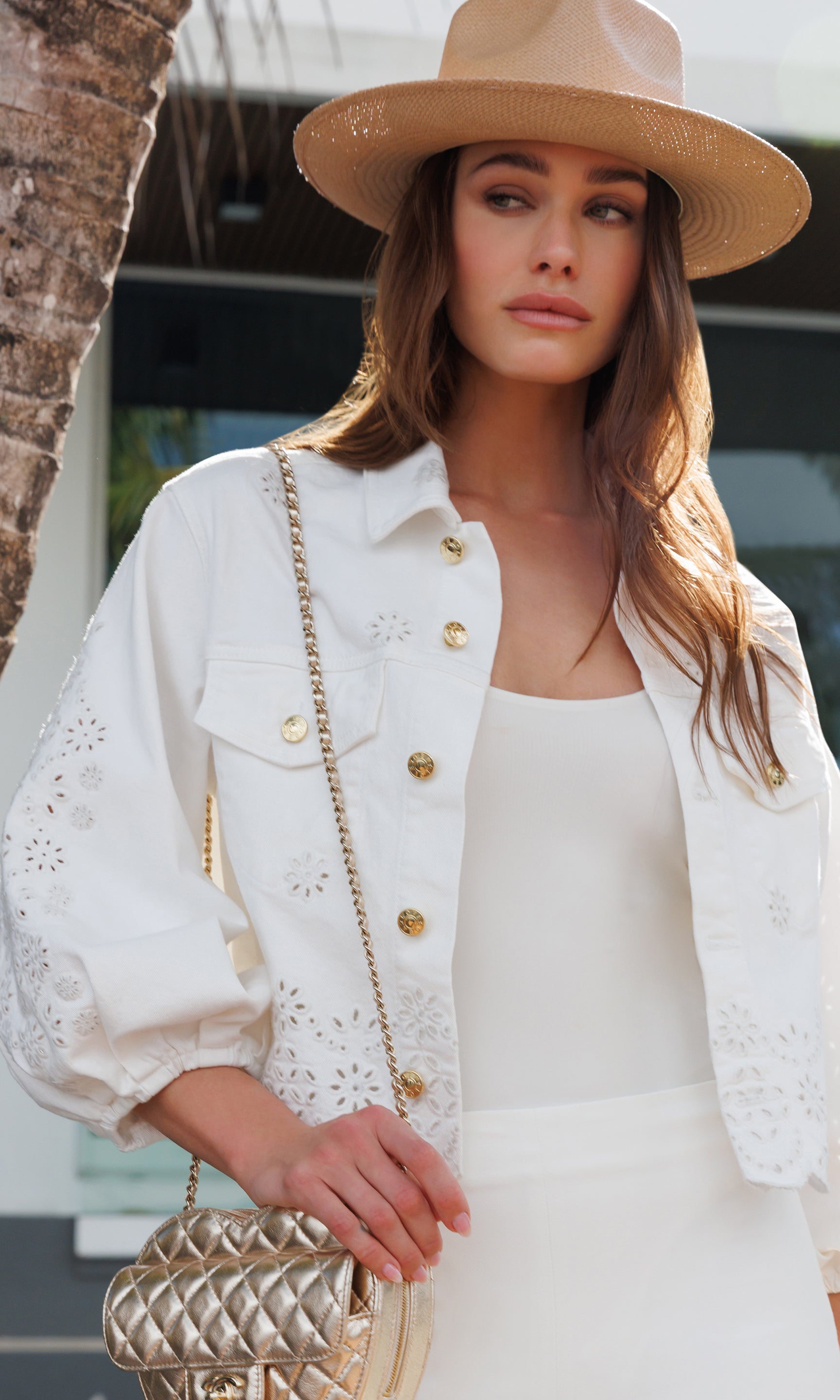 Woman wearing a white outfit with a beige hat and holding a gold handbag.