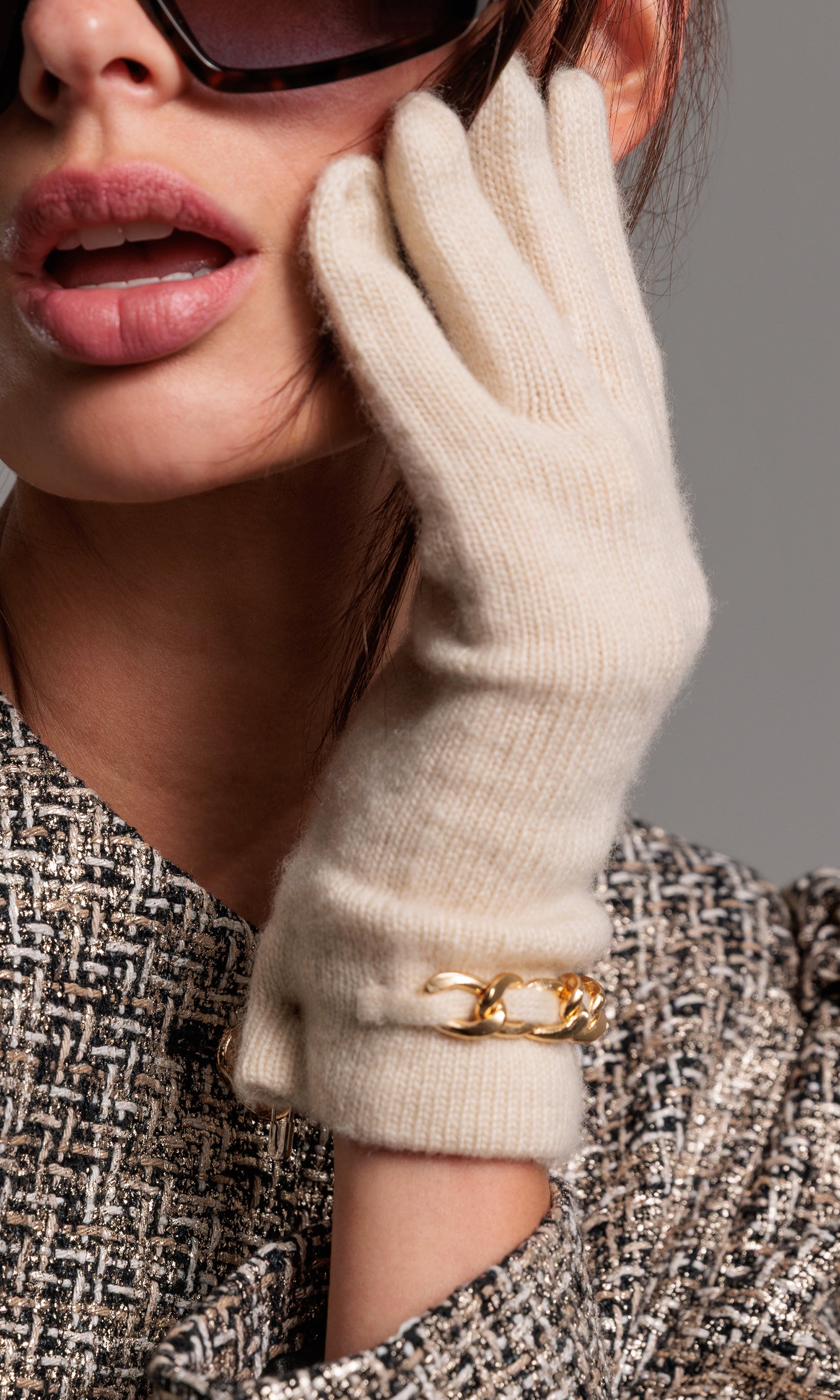 Close-up of a person wearing sunglasses, a beige glove, and a gold bracelet on a gray background