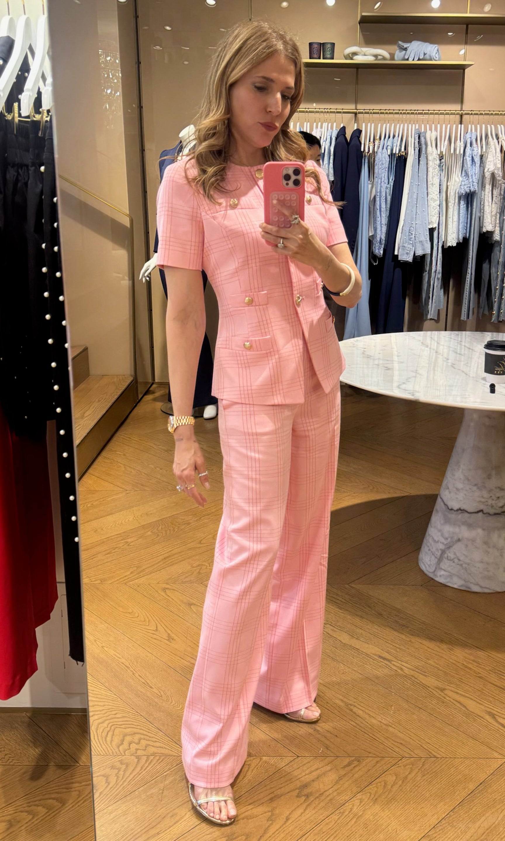 Woman taking a mirror selfie wearing a pink plaid suit in a clothing store.