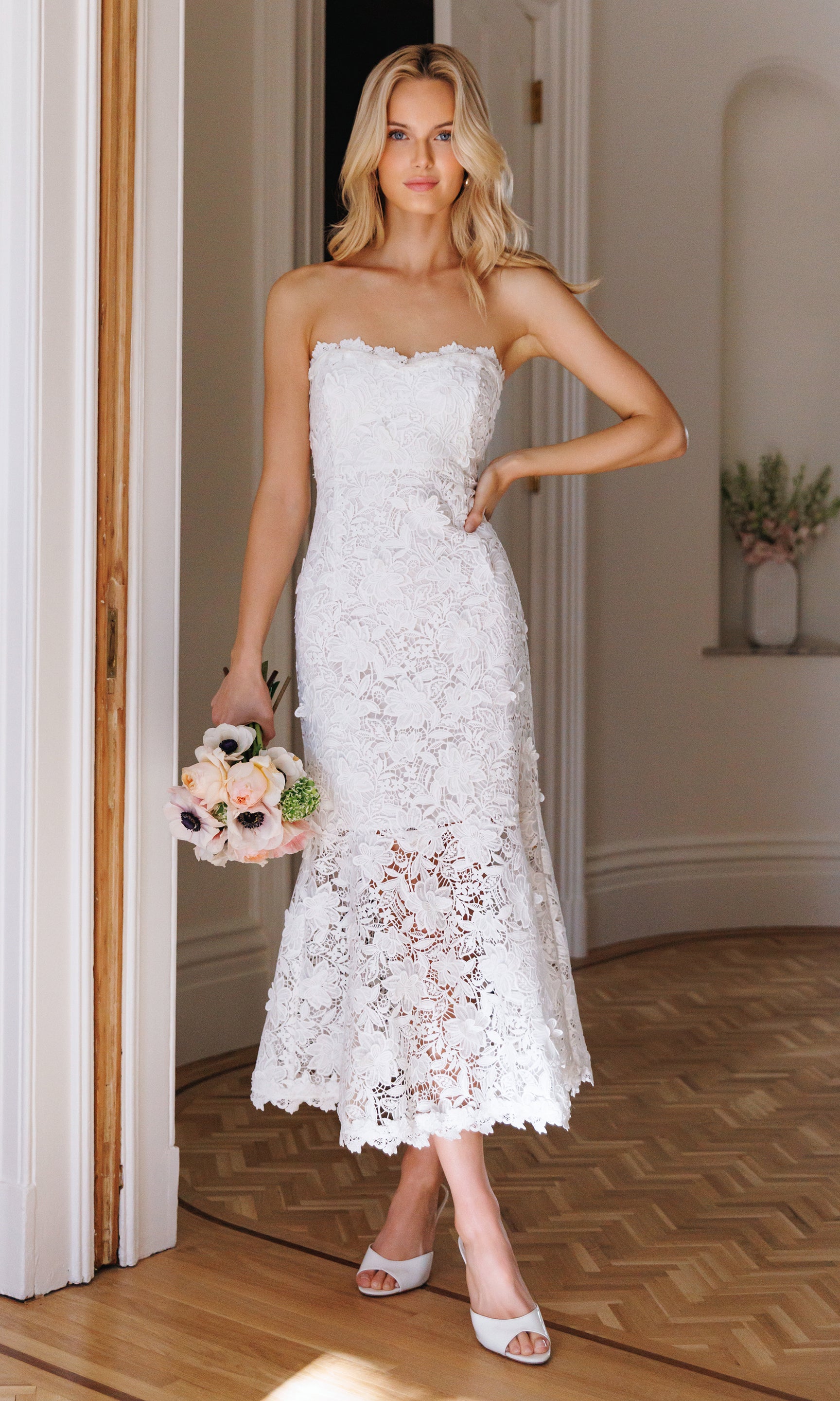 Woman in a white lace dress holding a bouquet indoors.