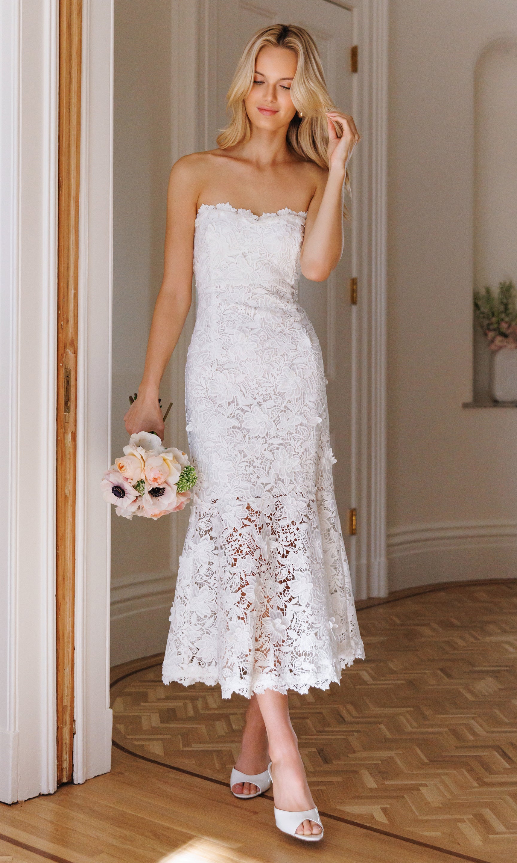 Woman in a white lace dress holding a bouquet in a room with wooden flooring.
