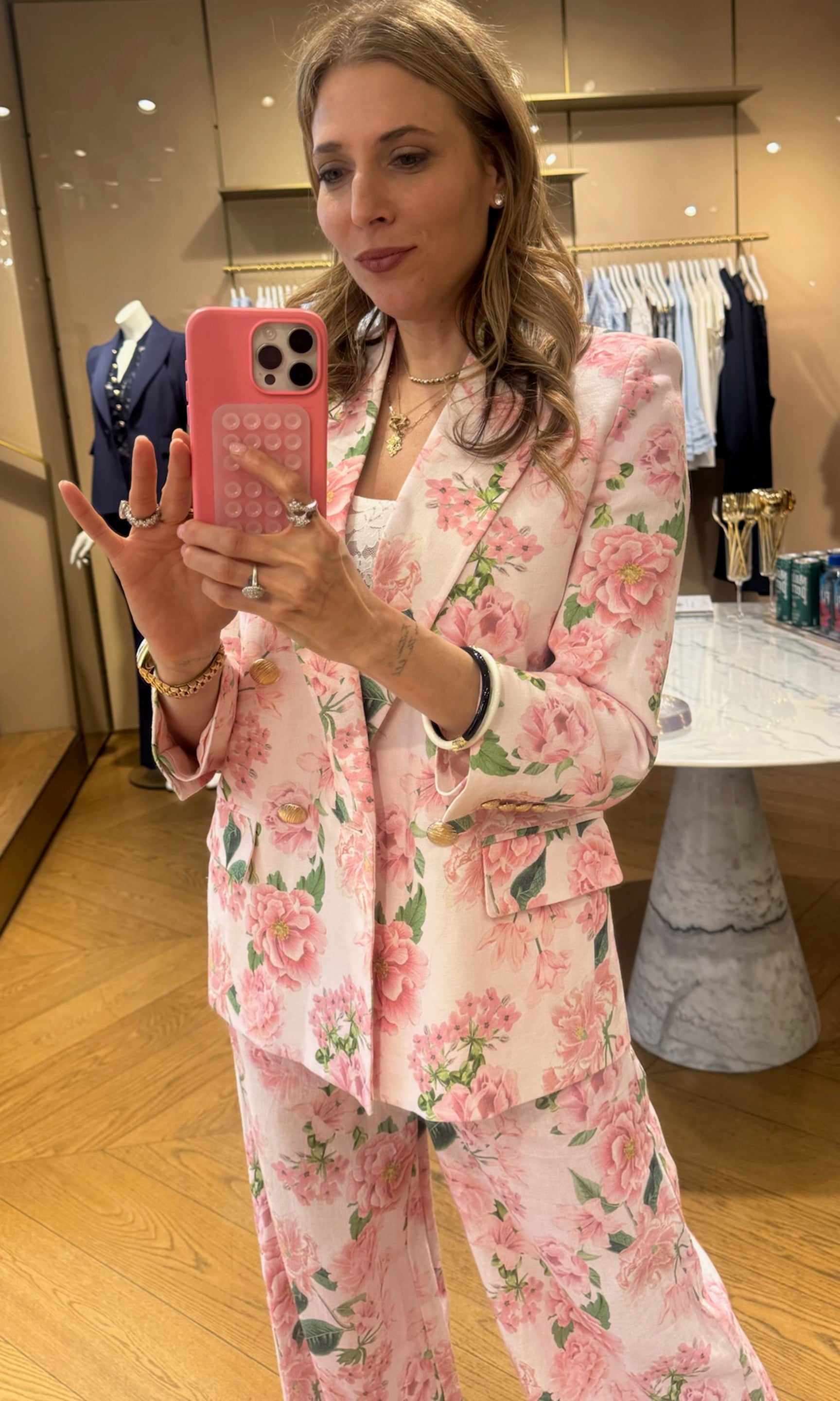 Roni taking a mirror selfie wearing a pink floral suit in a clothing store.