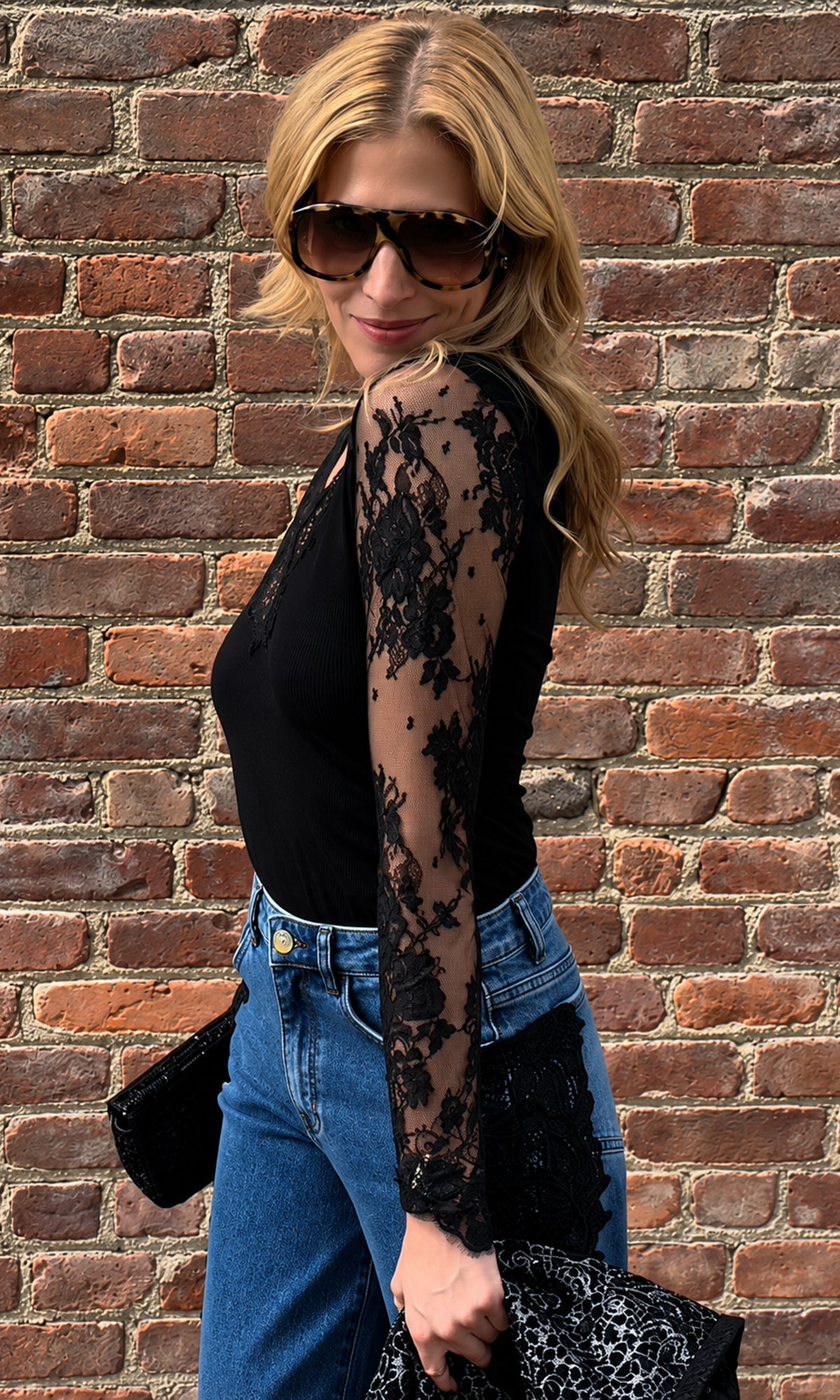 Our founder, Roni wearing a black long-sleeve top with sheer black lace sleeves, paired with blue jeans and sunglasses, standing against a red brick wall.