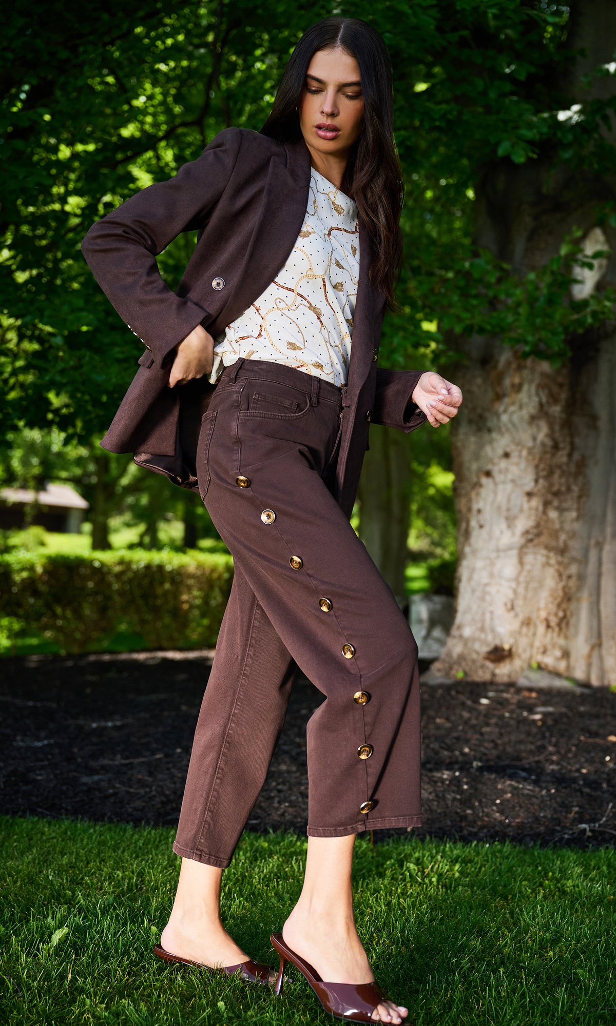 A woman posing outdoors in a park setting, wearing a dark brown blazer, matching brown buttoned pants, a white patterned top, and brown mule sandals.