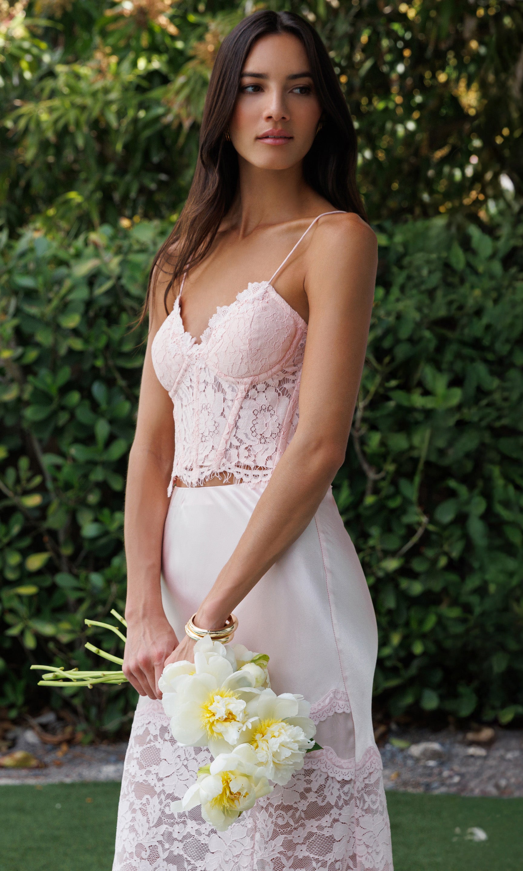Woman in a lace bustier and matching skirt holding flowers against a green foliage background