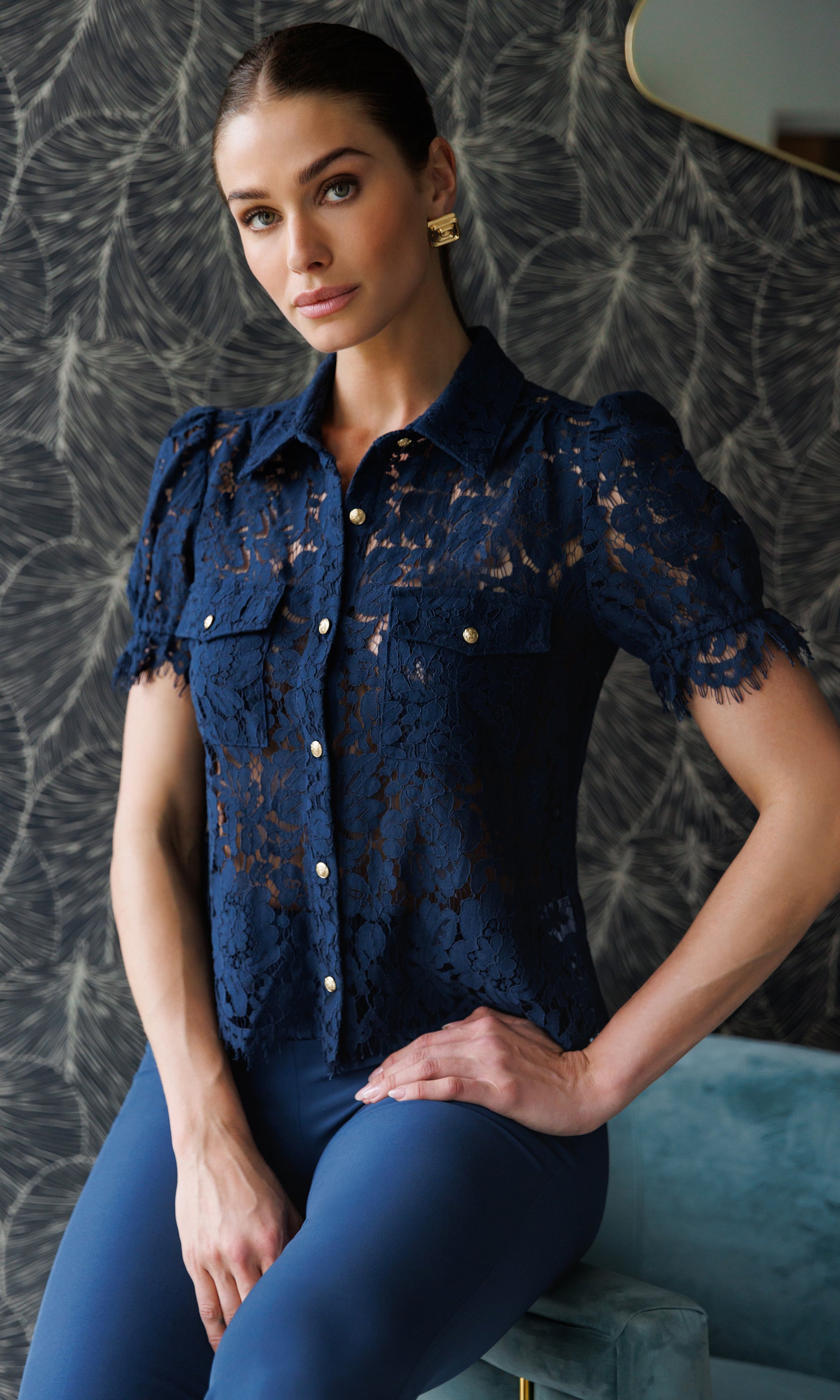 Woman wearing a navy lace top and blue pants against a textured wall.