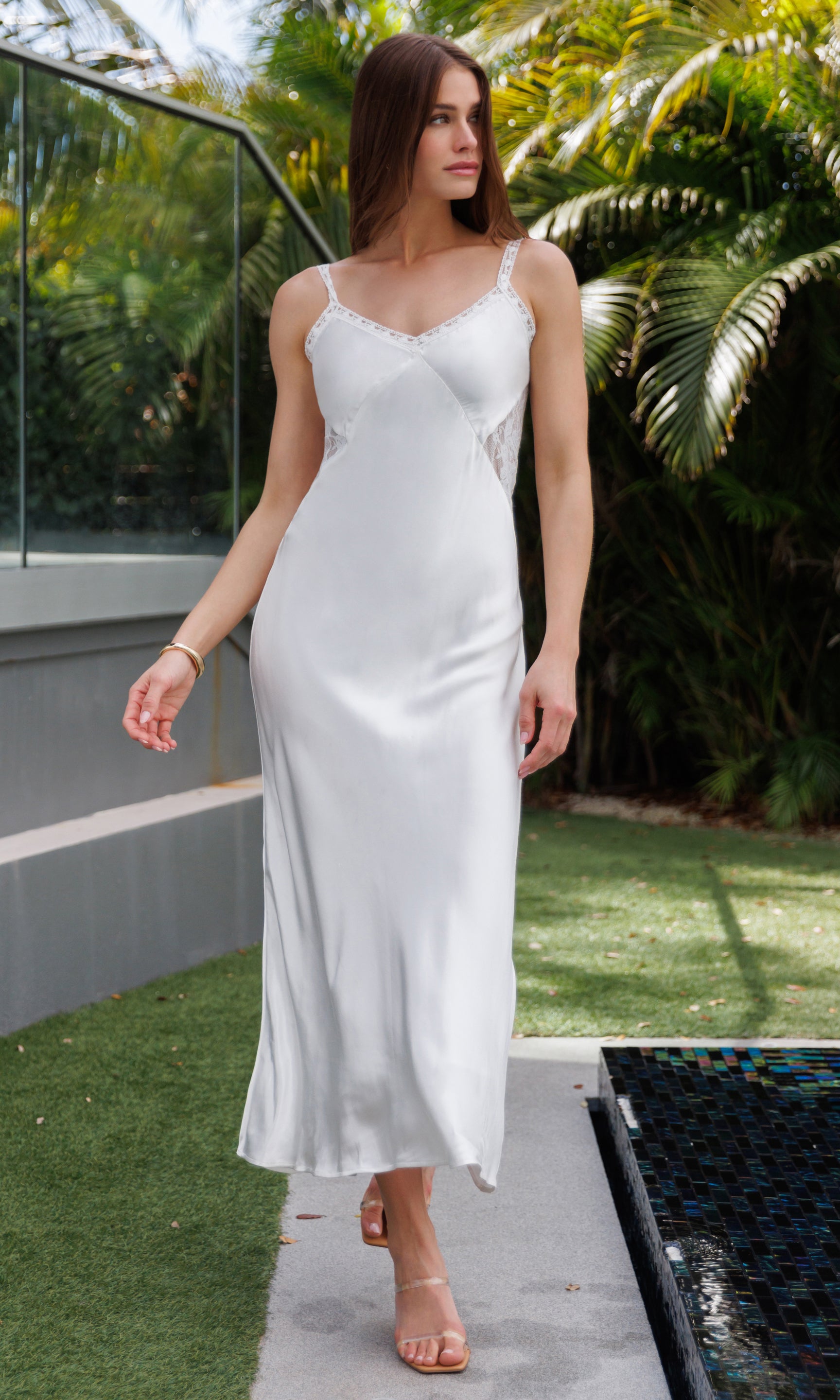 Woman in a white satin dress with lace details standing outdoors with greenery in the background