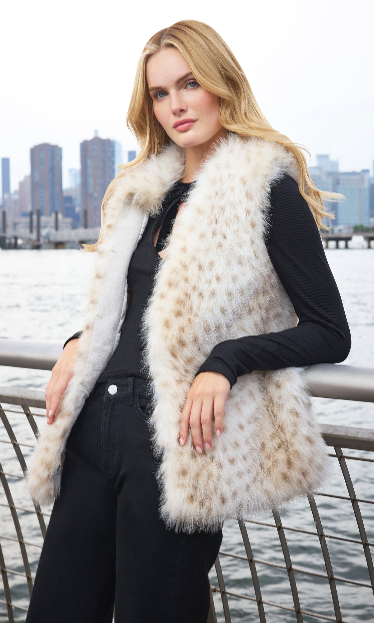 Model wearing a cream and tan faux fur vest over a black long-sleeve top and black pants, posing outdoors by a waterfront railing with a city skyline in the background.