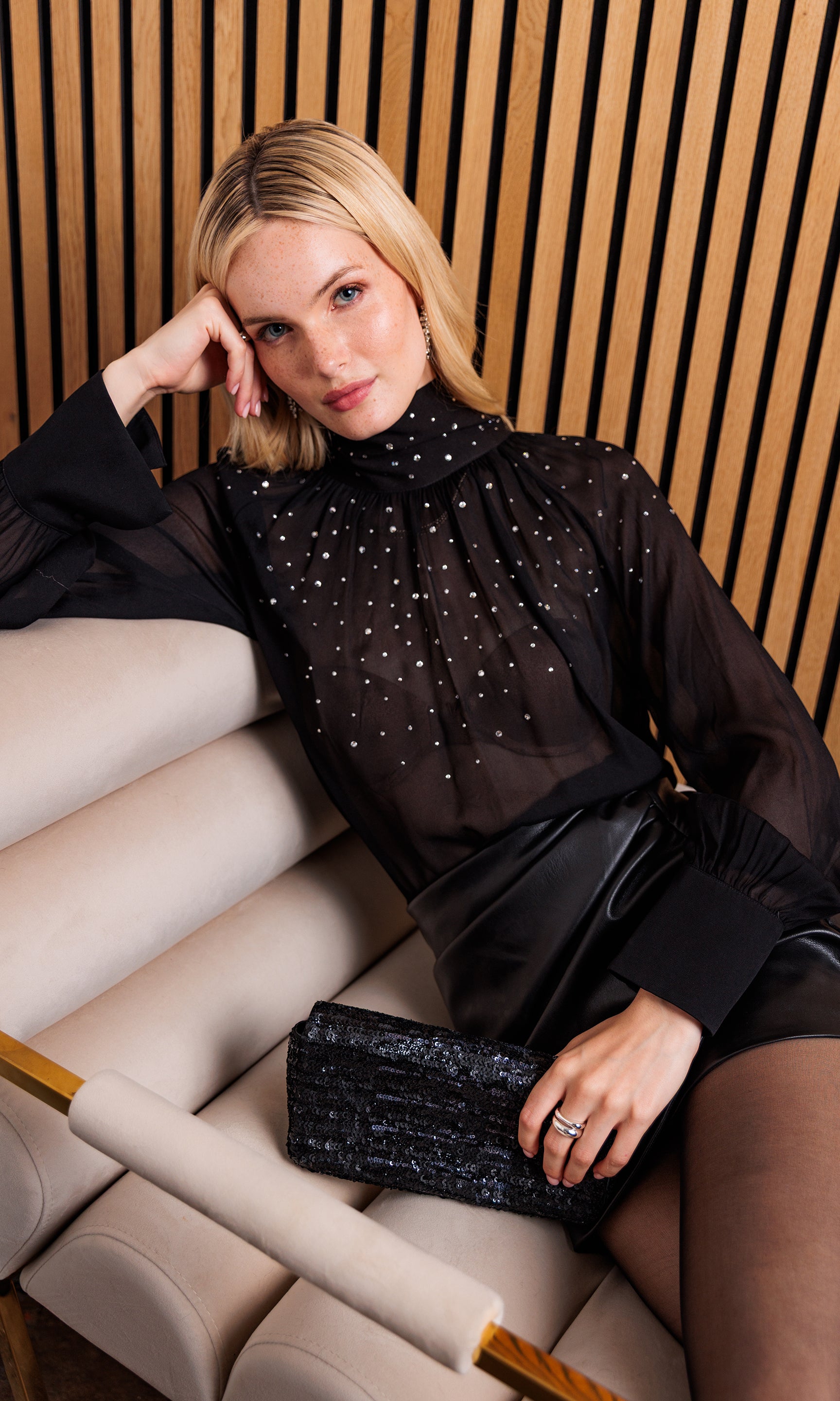 The model lounges in the Noor Crystal Blouse, showing a relaxed pose that highlights the blouse’s sheer elegance and crystal accents.