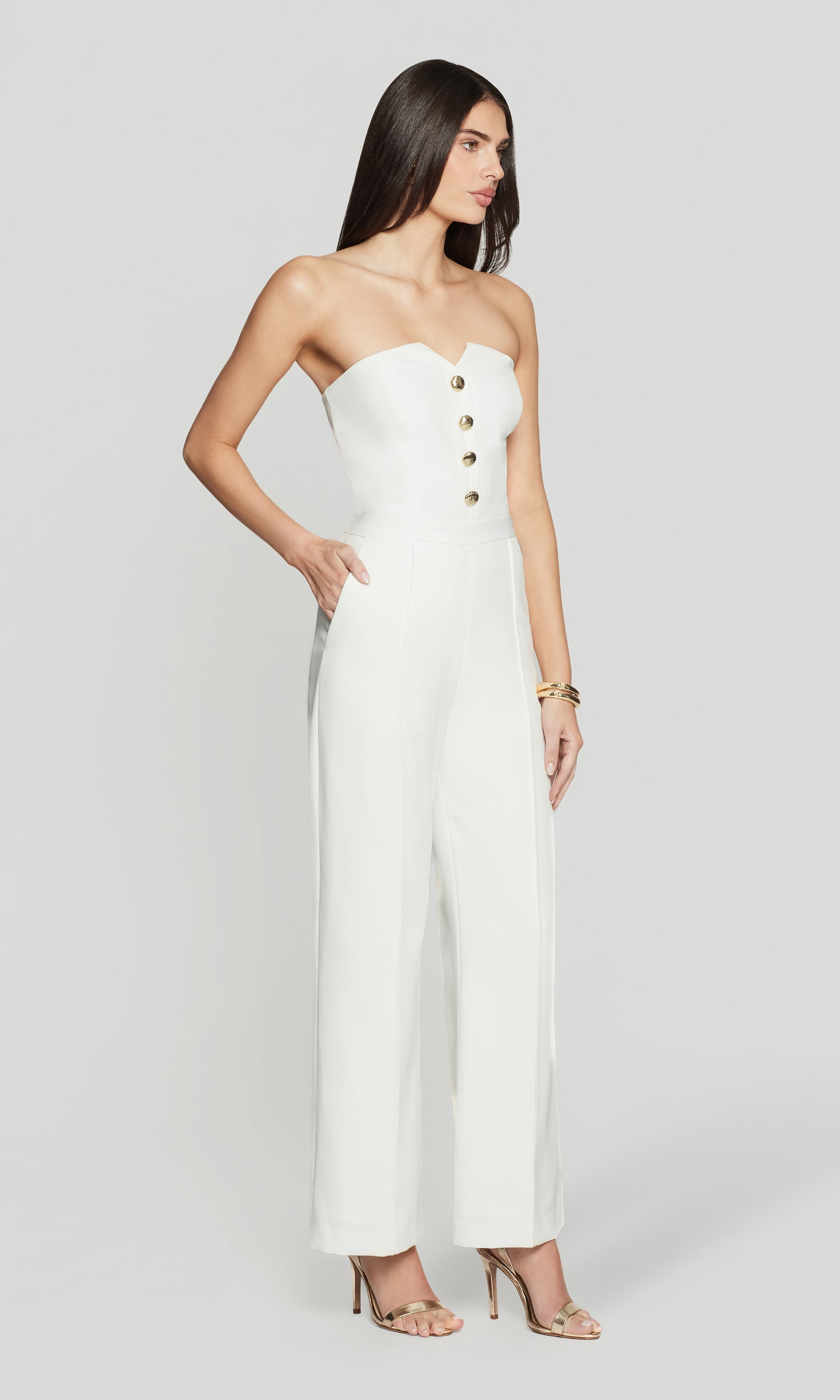 Side view of woman wearing a strapless white jumpsuit with gold buttons on a white background