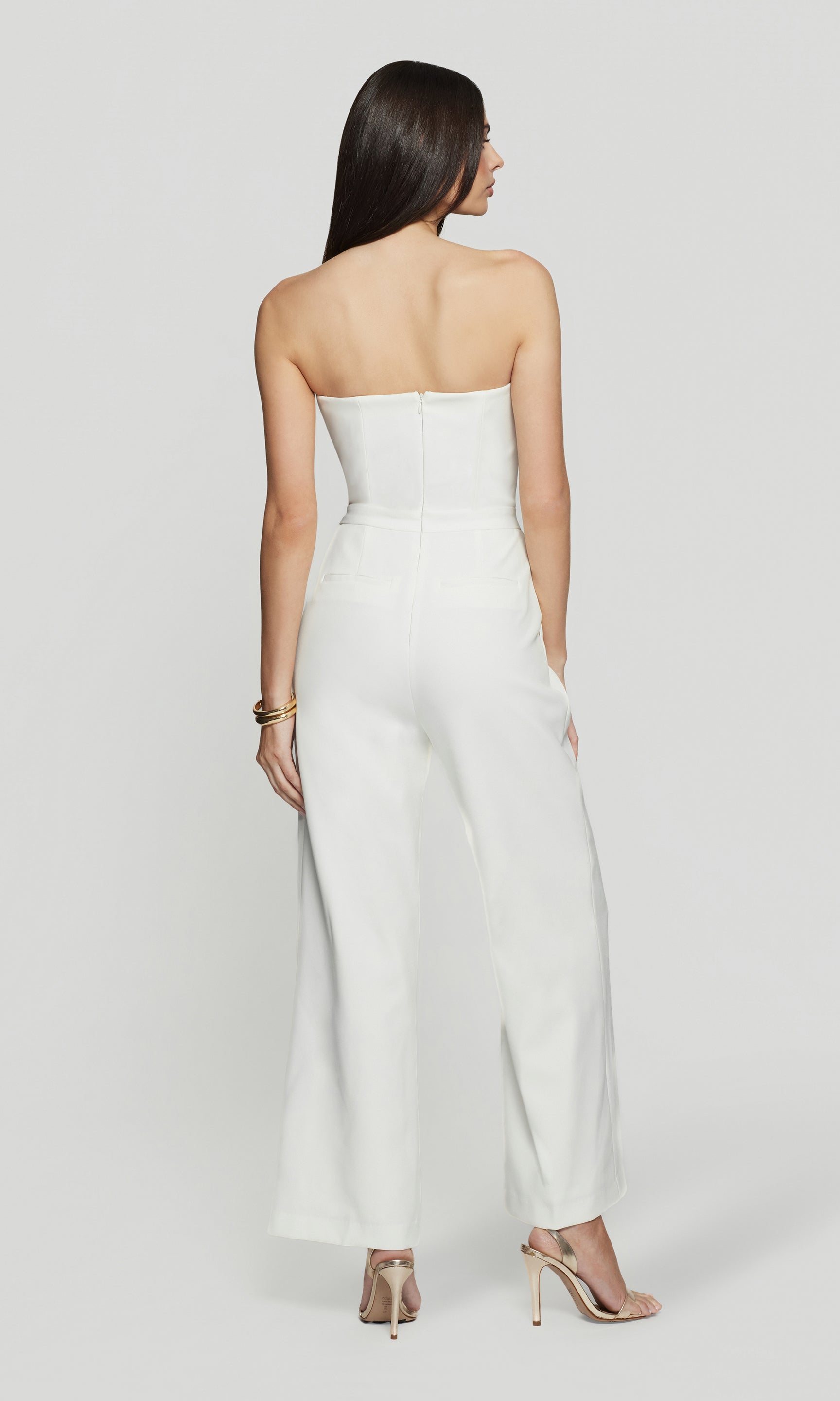 Back view of woman wearing a strapless white jumpsuit on a plain background