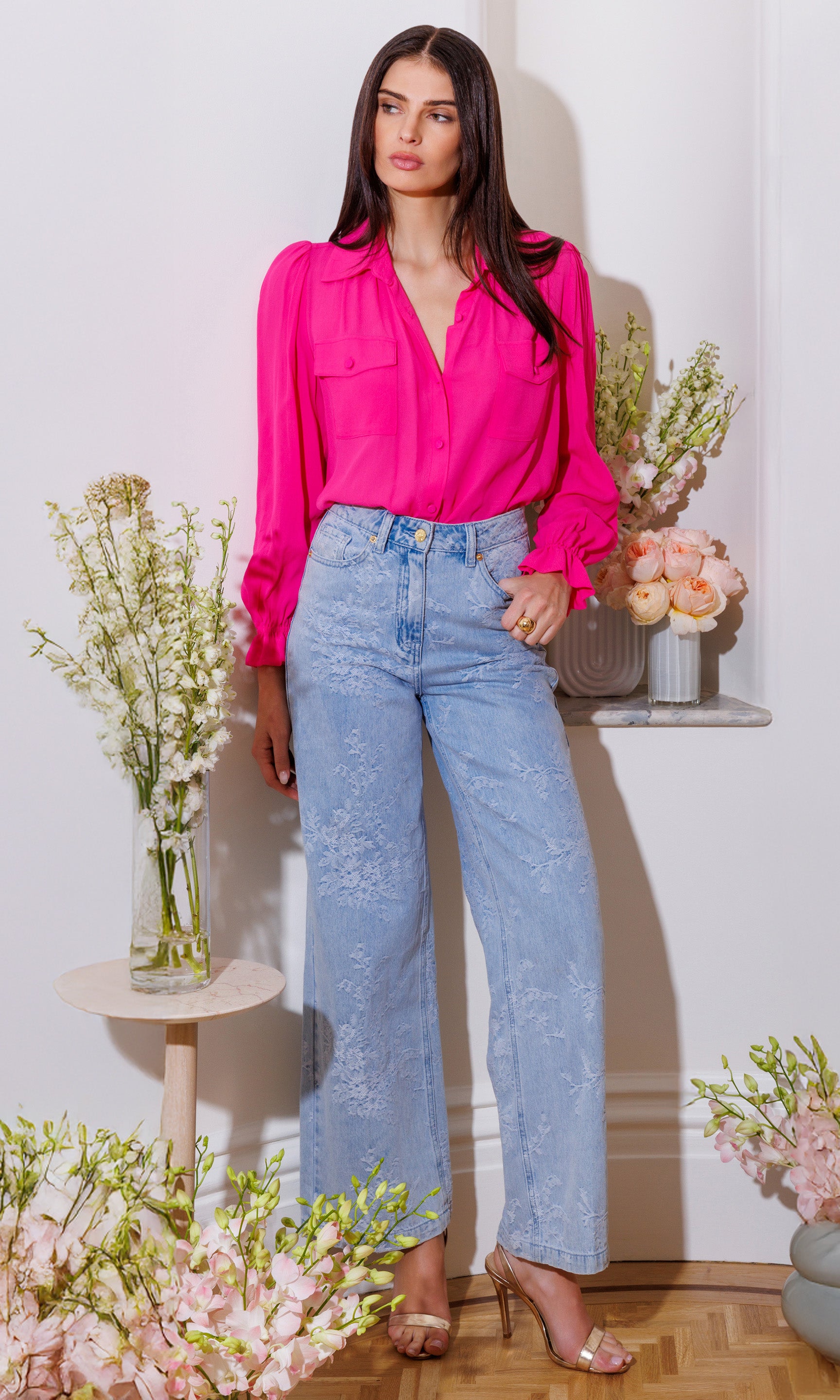 Woman wearing a bright pink blouse and light blue jeans standing in a room with decorative flowers and a small table.