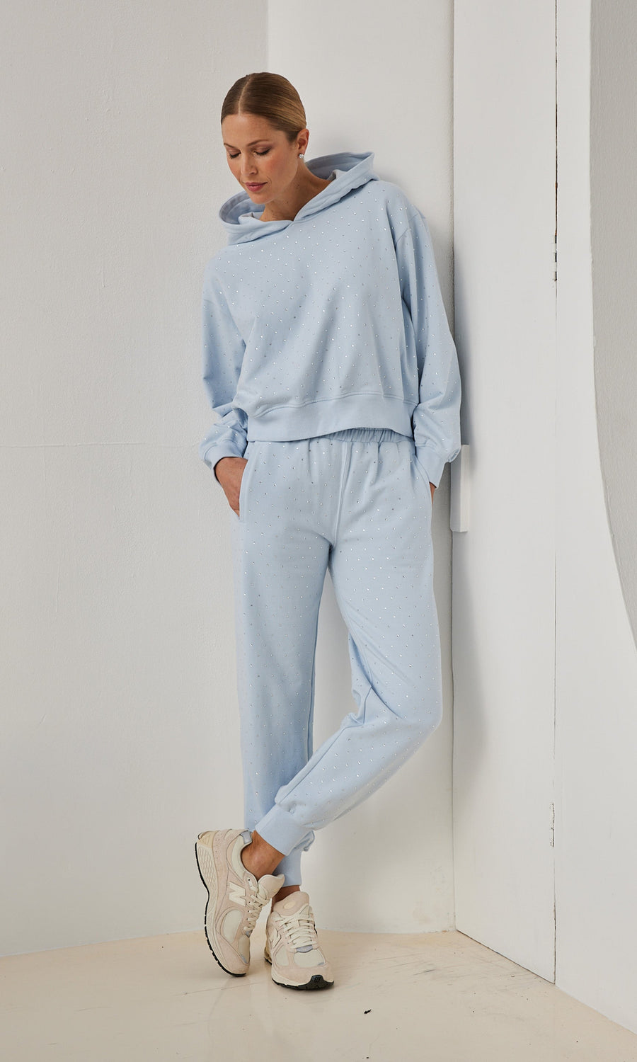 A model in a light blue sweatshirt and sweatpants strikes a casual pose, leaning slightly to one side with a friendly expression.