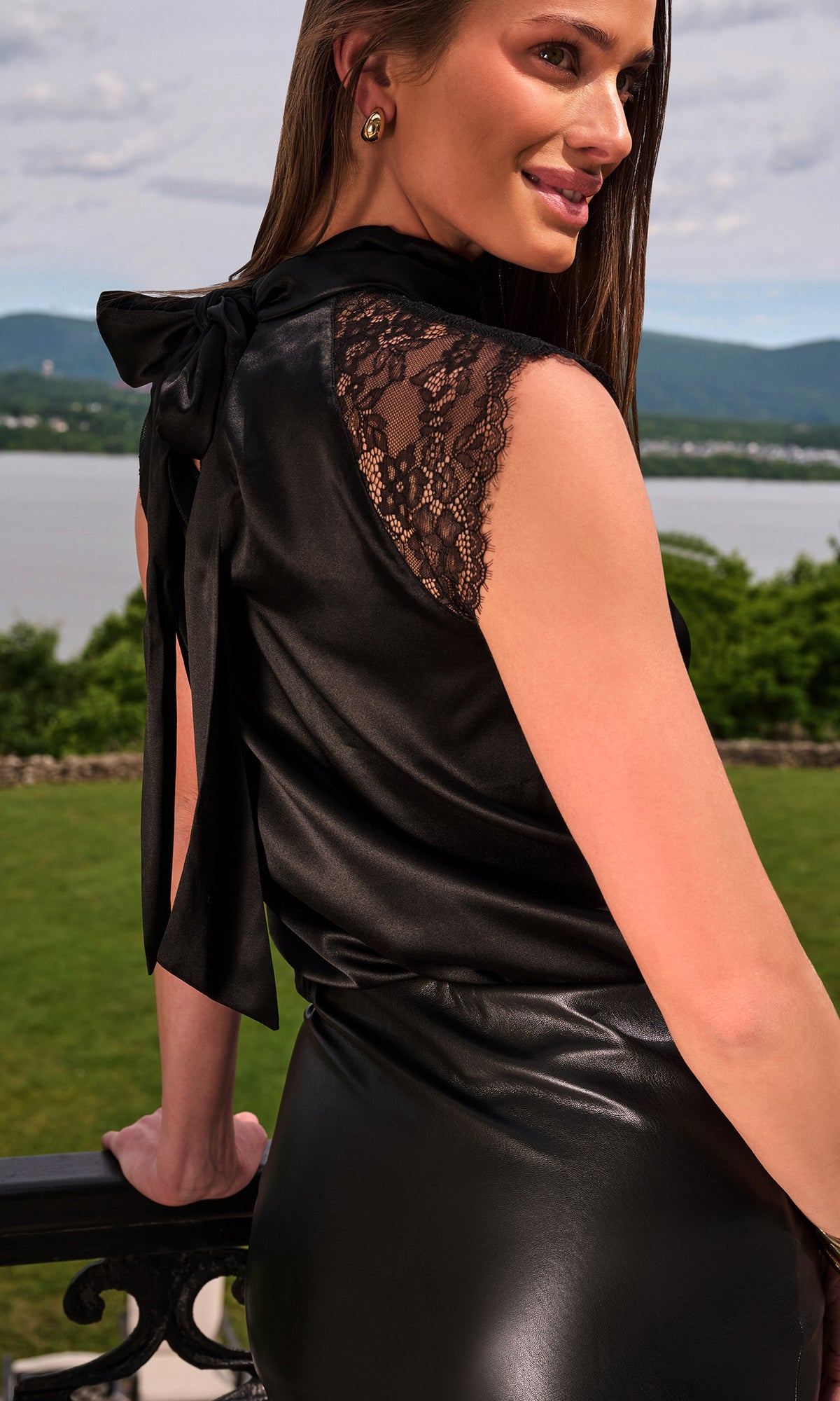 Model wearing a black satin-like sleeveless dress with a large bow at the back of the neck and lace shoulder detailing.