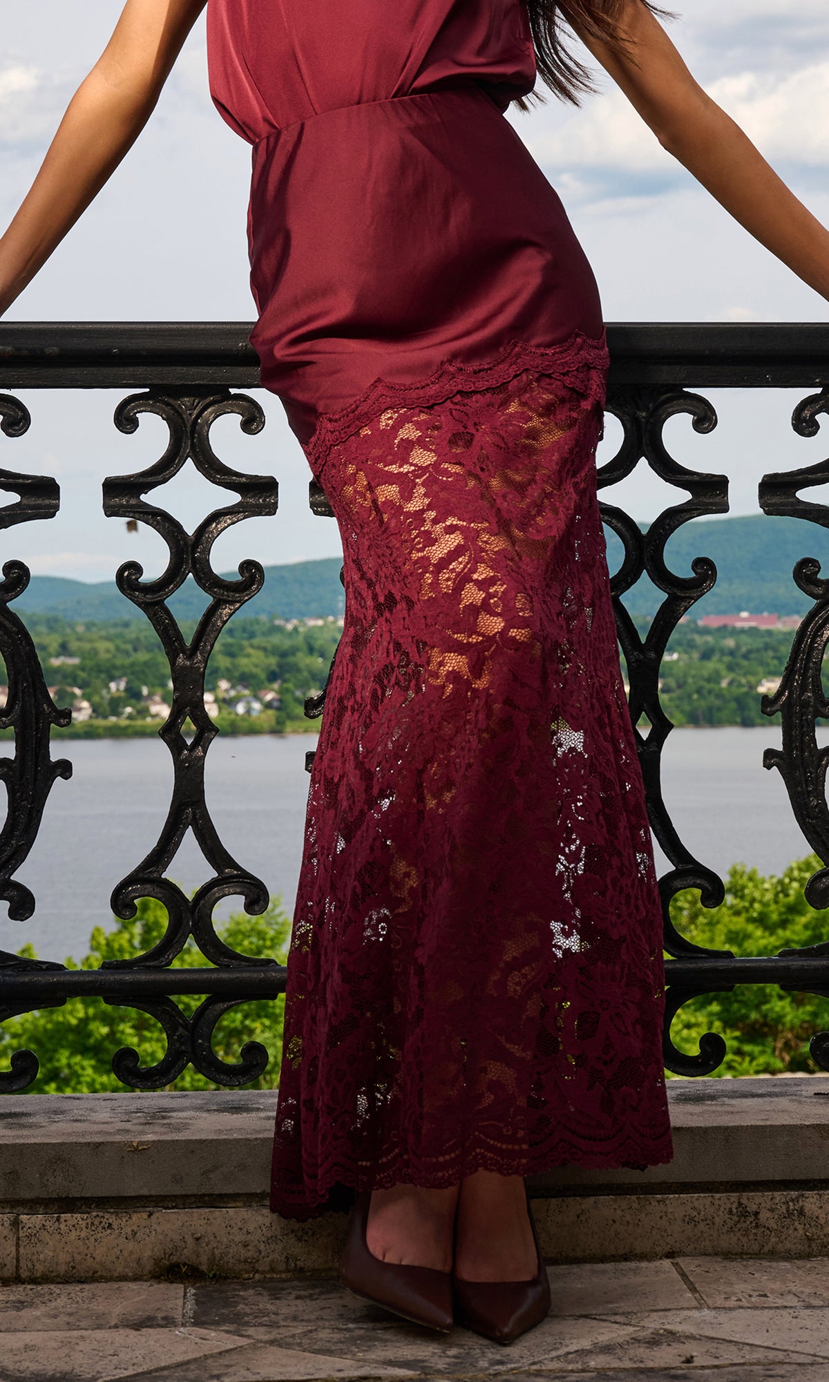 A woman wearing a long burgundy maxi skirt with a lace lower hem, standing on a stone terrace with a scenic lake and mountains in the background.