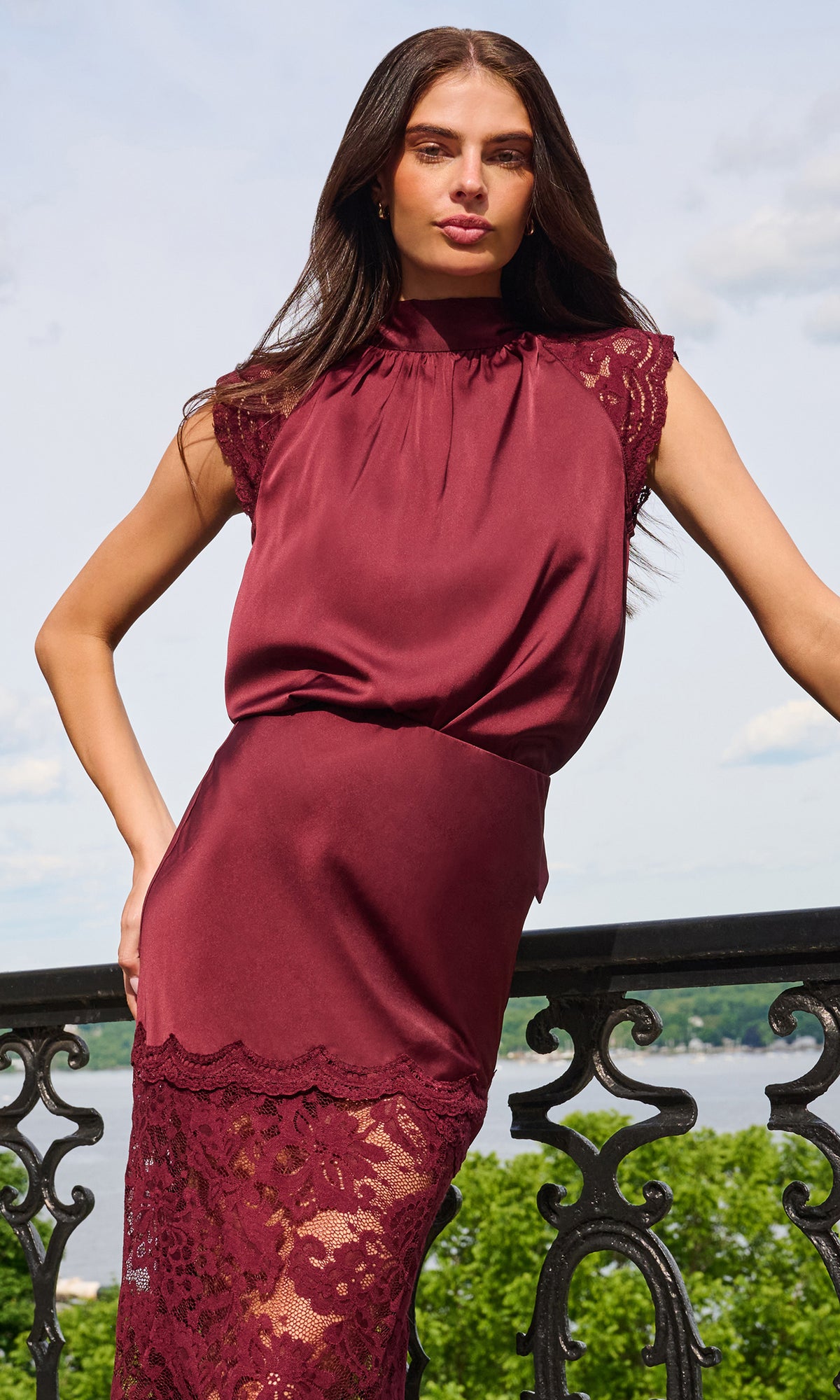 A woman models a sleeveless high-neck dress in deep burgundy with lace trim at the cap sleeves and hem, photographed outdoors against a railing and sky.