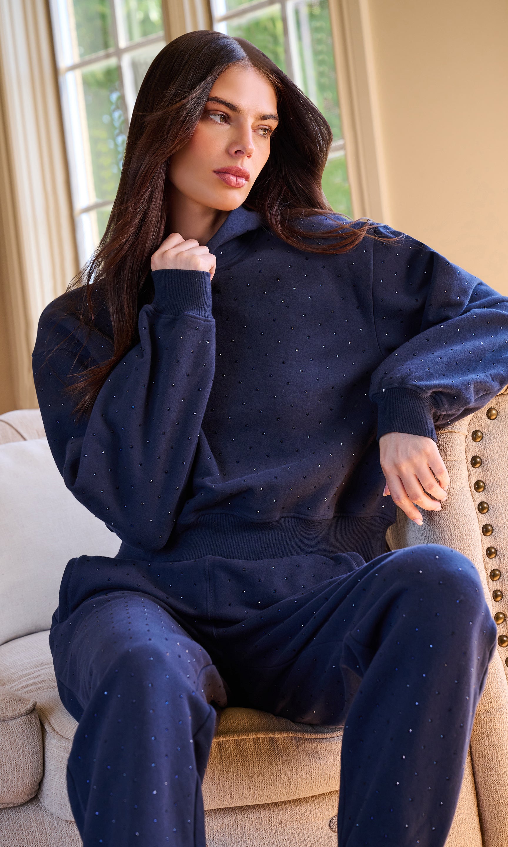 Model wearing a matching navy blue loungewear set with crystal embellishments , sitting on a beige armchair in a brightly lit room.
