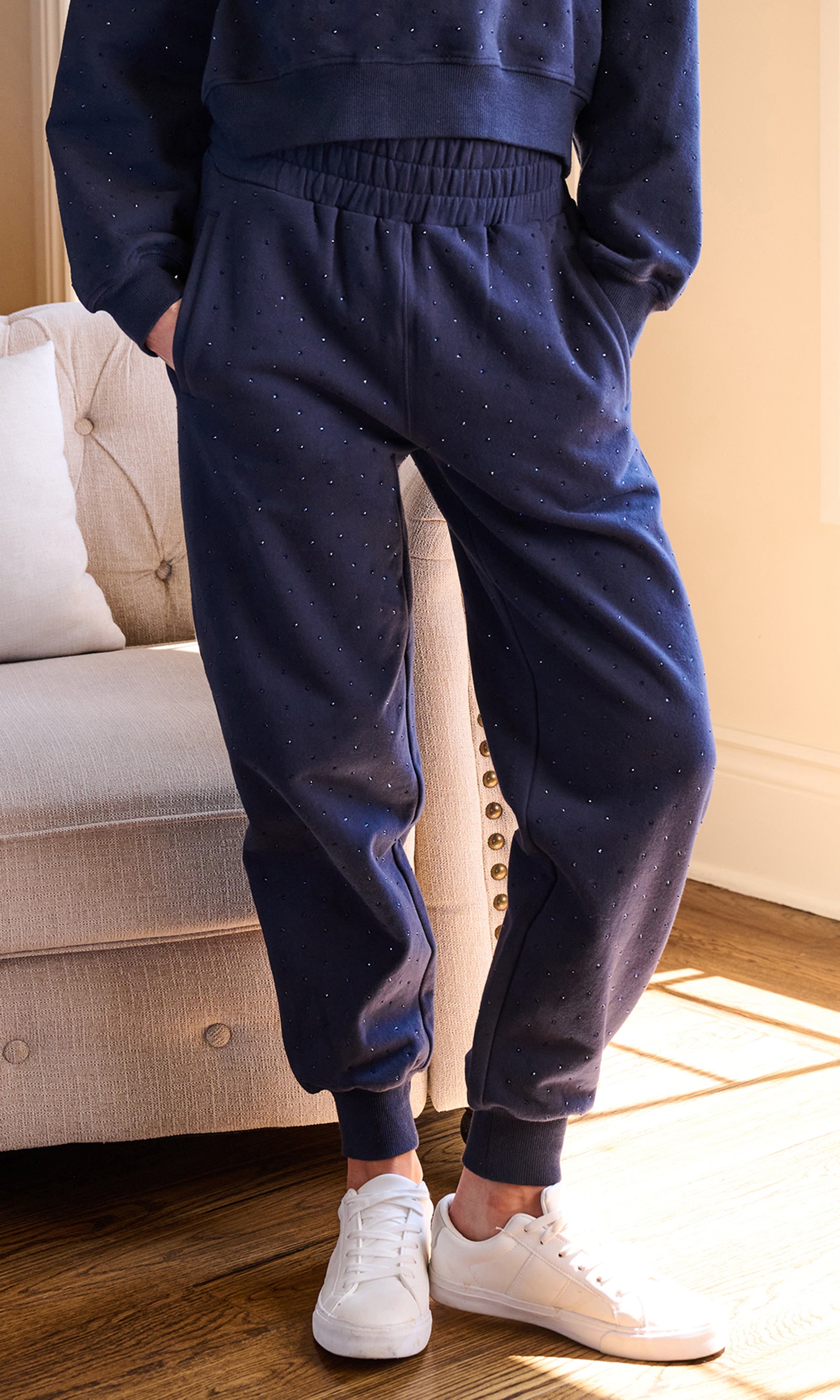 Model wearing navy blue jogger sweatpants with a subtle speckled pattern, paired with white sneakers, standing in a brightly lit room next to a beige sofa.