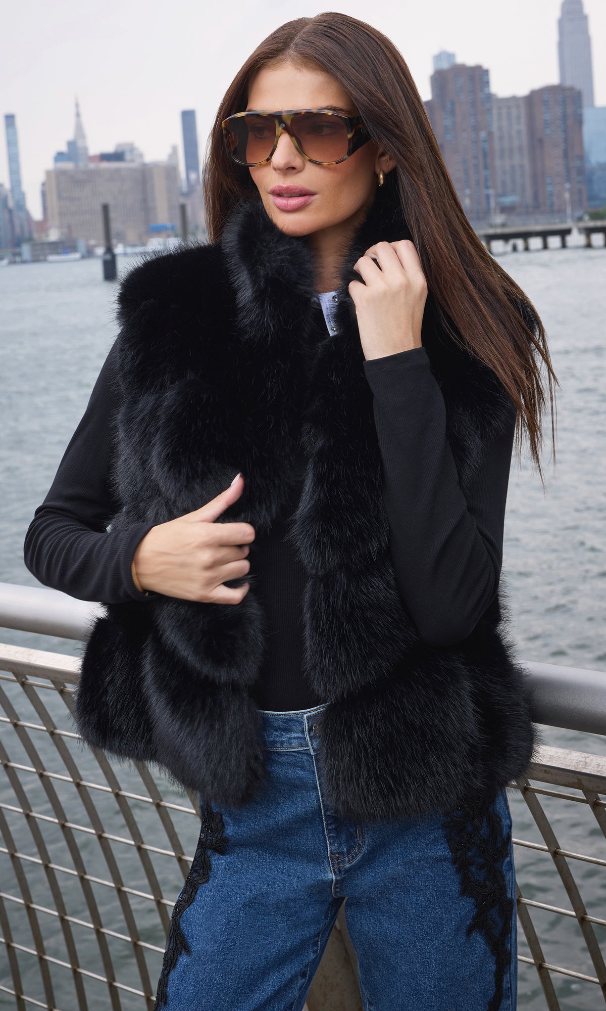 A woman wearing a black fur vest over a black long-sleeve top and blue jeans, standing outdoors near a waterfront railing with a city skyline in the background.