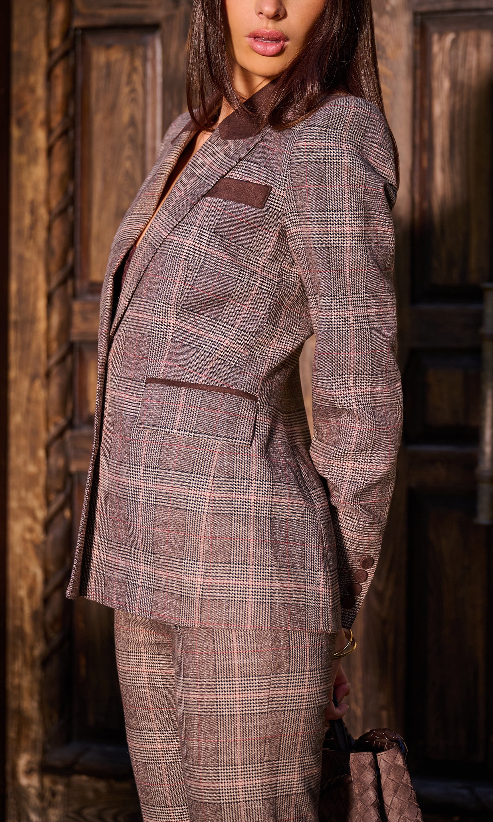 A woman wearing a tailored plaid suit in muted brown, beige, and gray tones, standing in front of a rustic wooden door. 