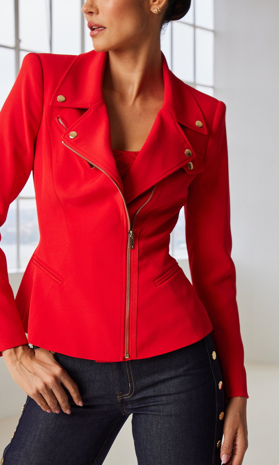 Model in a red moto jacket with gold details and jeans stands by a window.