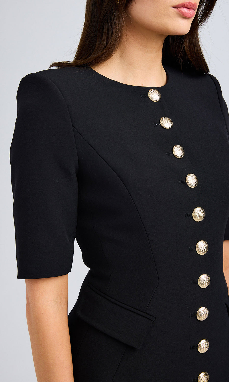 Close-up of a model in a black dress with front buttons, wearing a black crepe jacket with gold buttons and short sleeves.