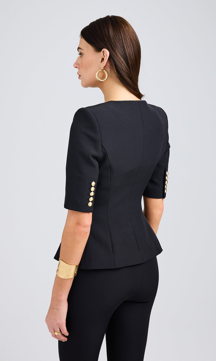 Woman's back in a black blazer with short sleeves and gold buttons, wearing black pants, showcasing a stylish look.