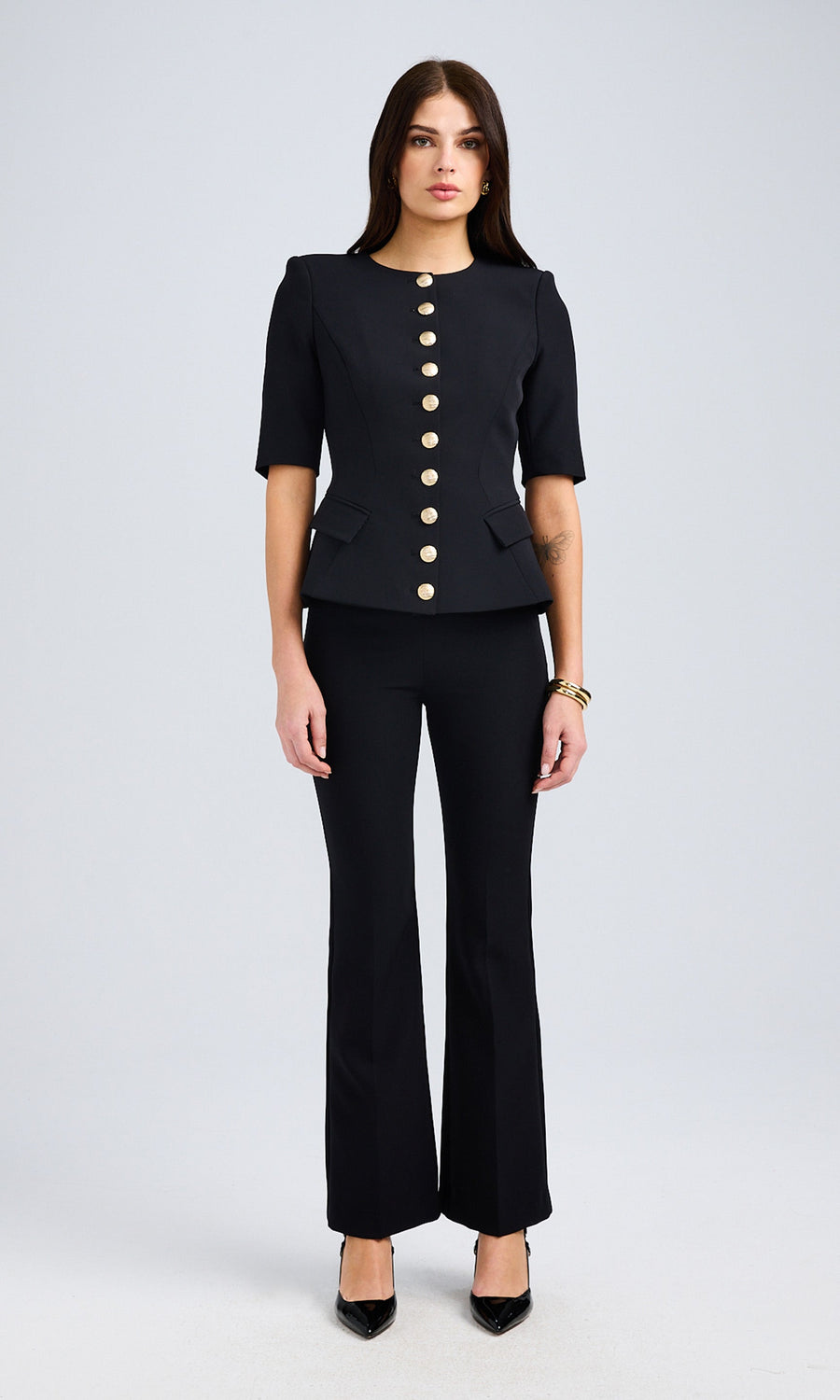 A model poses in a black crepe jacket with gold buttons and black pants, showcasing a stylish and elegant look.