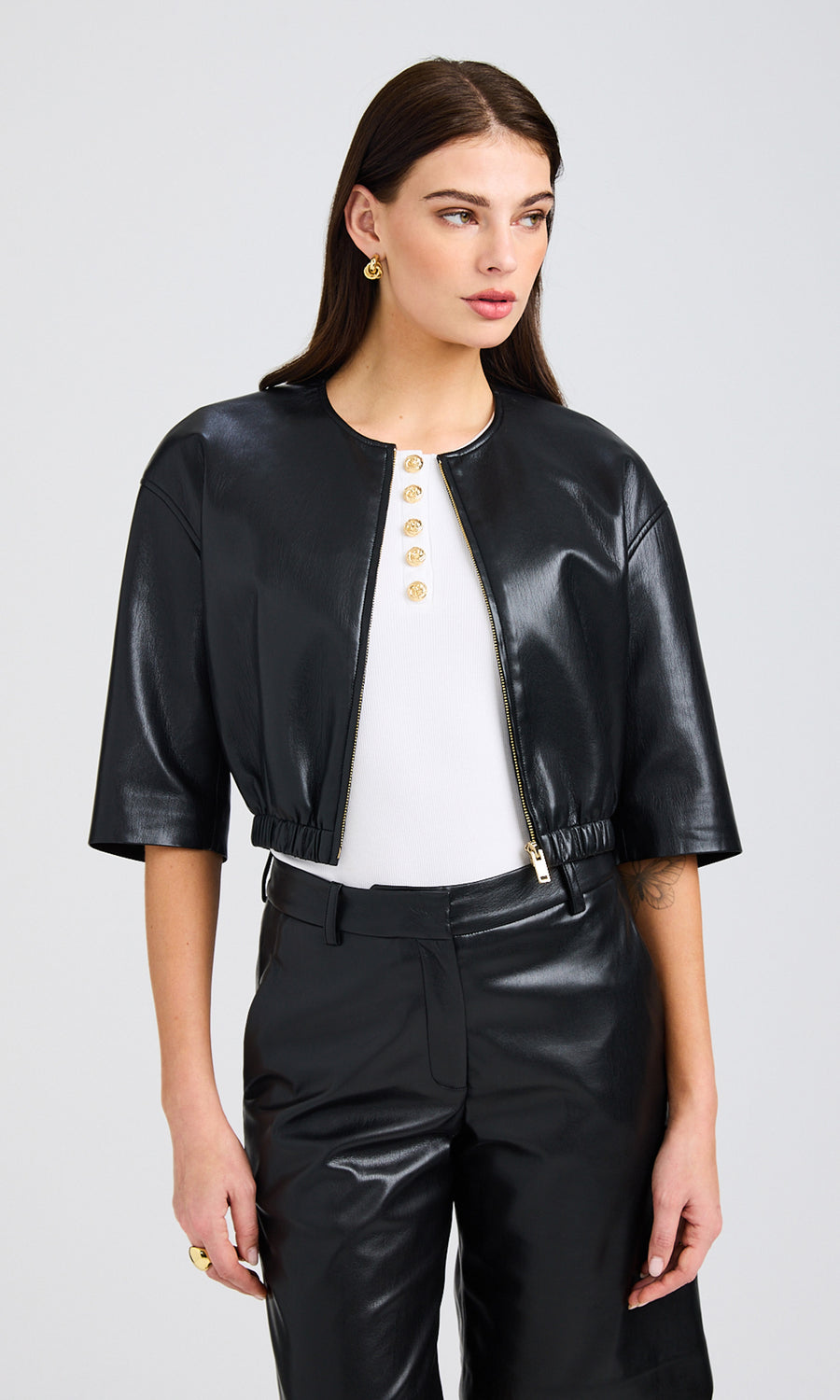 A Front view of the model wearing the vegan leather bomber, styled over a white top with gold button accents, paired with black vegan leather shorts.