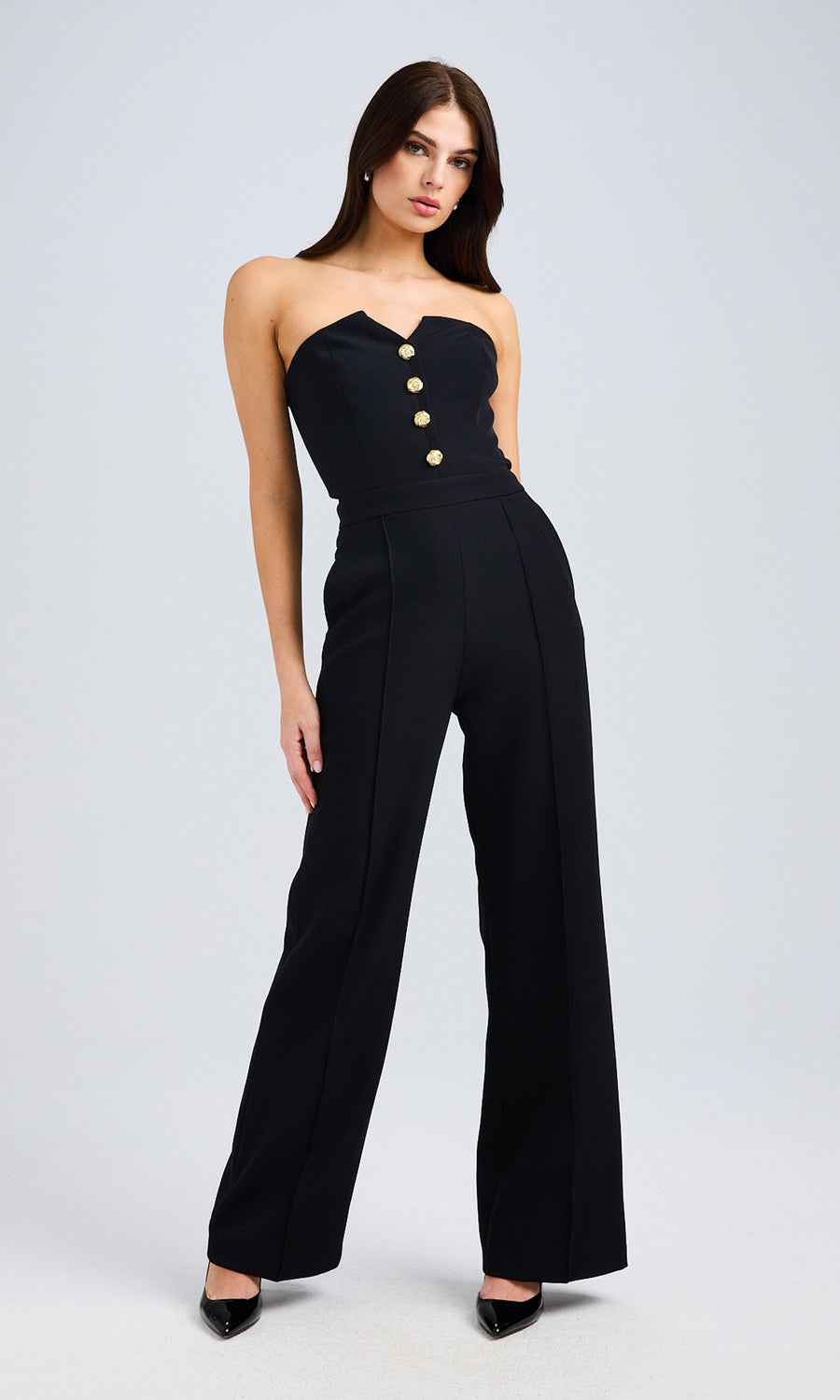 Full-body front view of the model in a black strapless jumpsuit with hands in pockets, styled with pointed black heels.