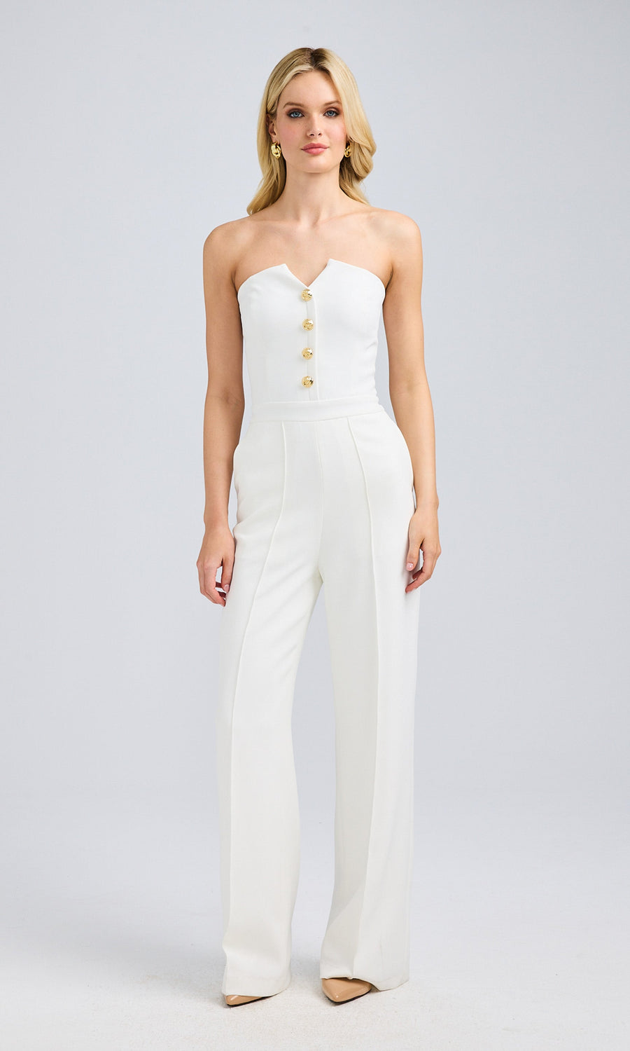 Front view of a blonde model in a white strapless jumpsuit with gold button details, posing with one hand relaxed at her side.