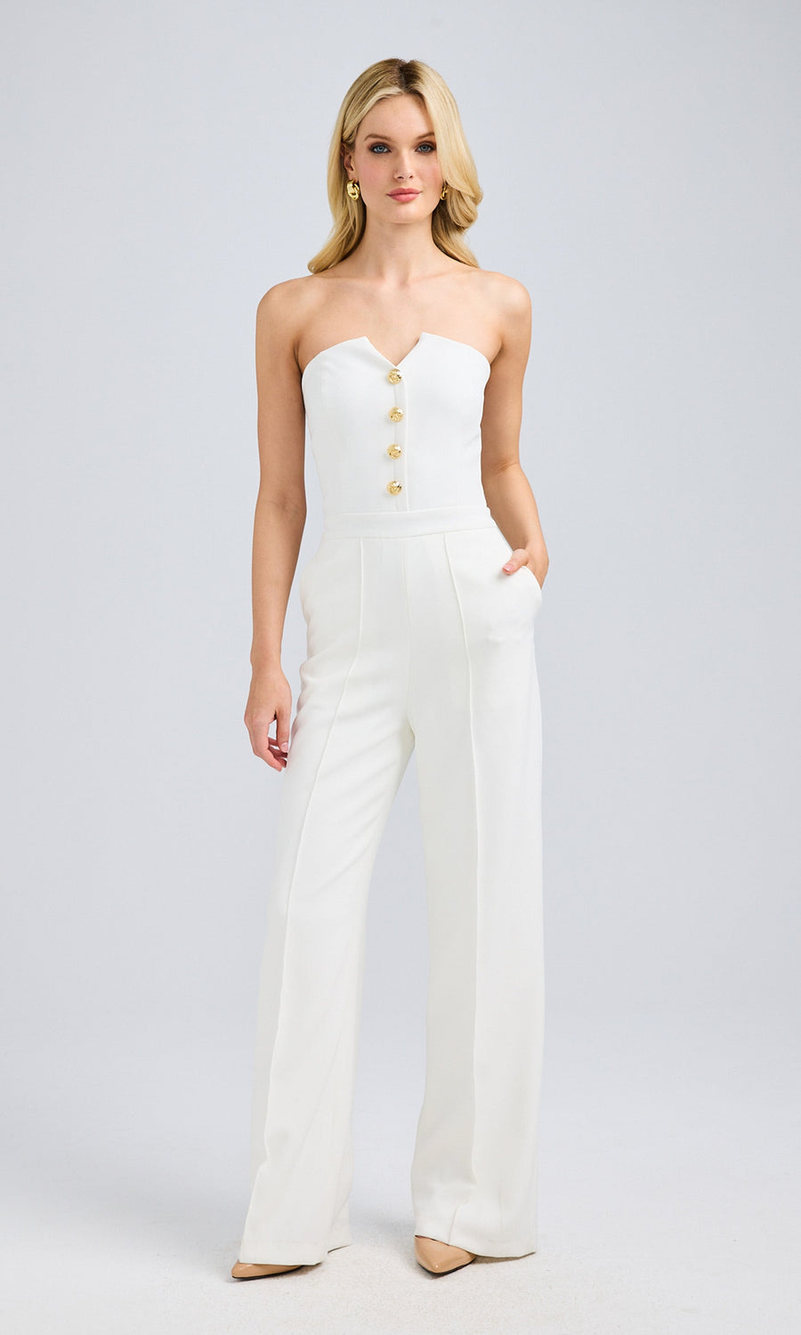 Full-length view of a blonde model in a white strapless jumpsuit, hands in pockets, paired with beige pointed-toe heels.