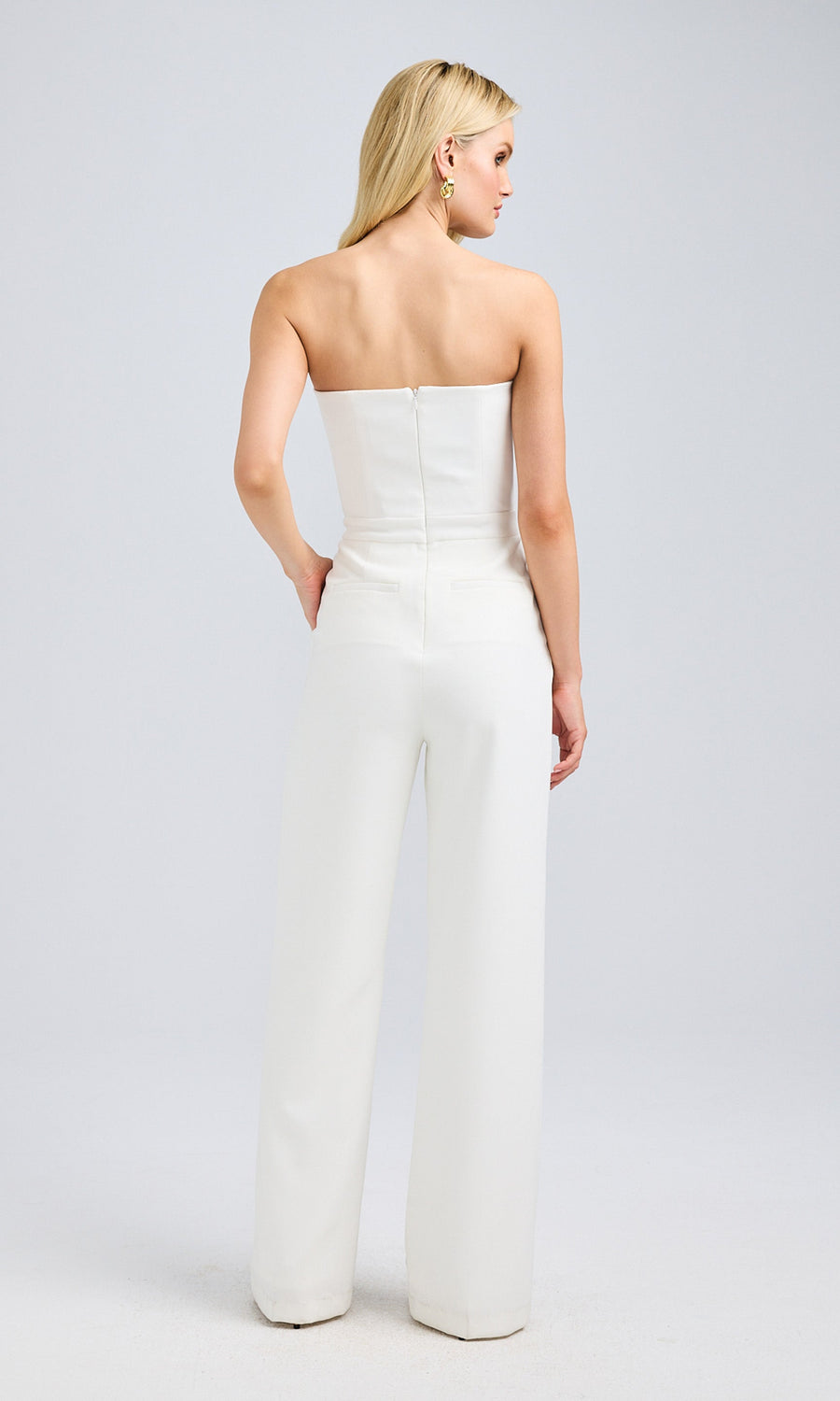 Back view of a white strapless jumpsuit, showcasing a zipper closure and tailored fit, worn by a blonde model