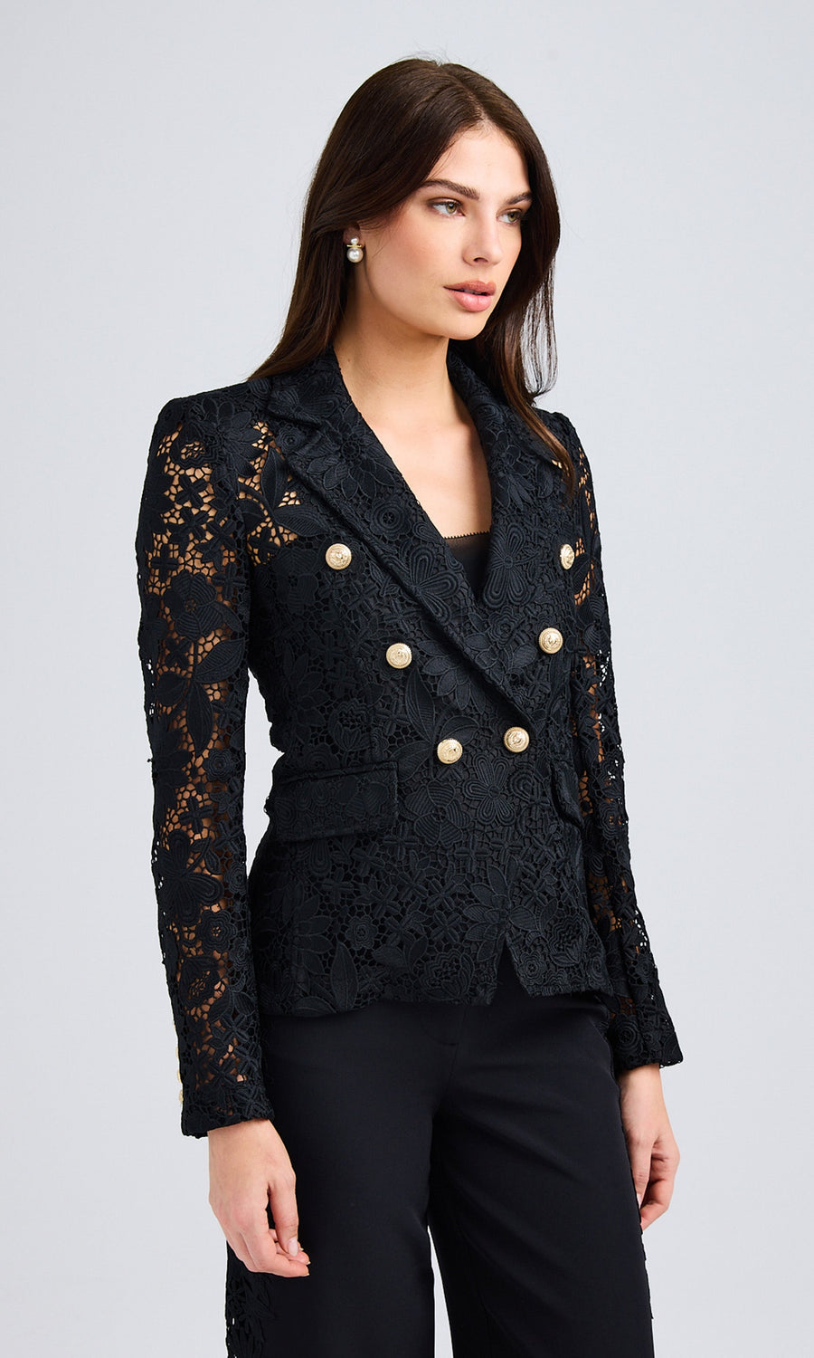 Front view of the model wearing a black lace blazer with gold button accents, styled with black trousers.