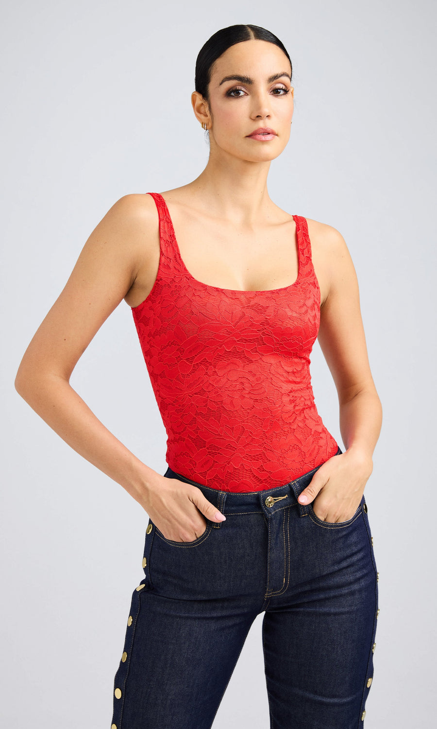 A woman wearing a red lace bodysuit and jeans stands confidently, showcasing her stylish outfit.