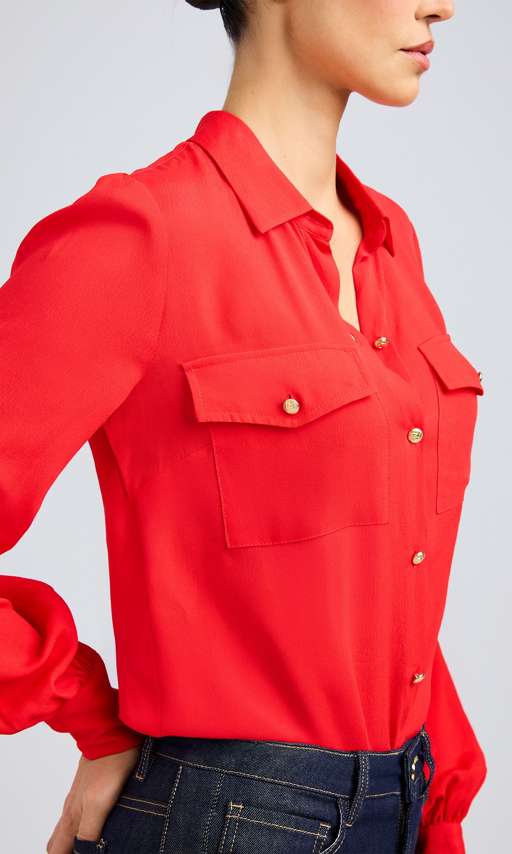 Close-up side profile of a flame red blouse with pleated sleeves and gold-tone buttons, paired with dark denim pants.