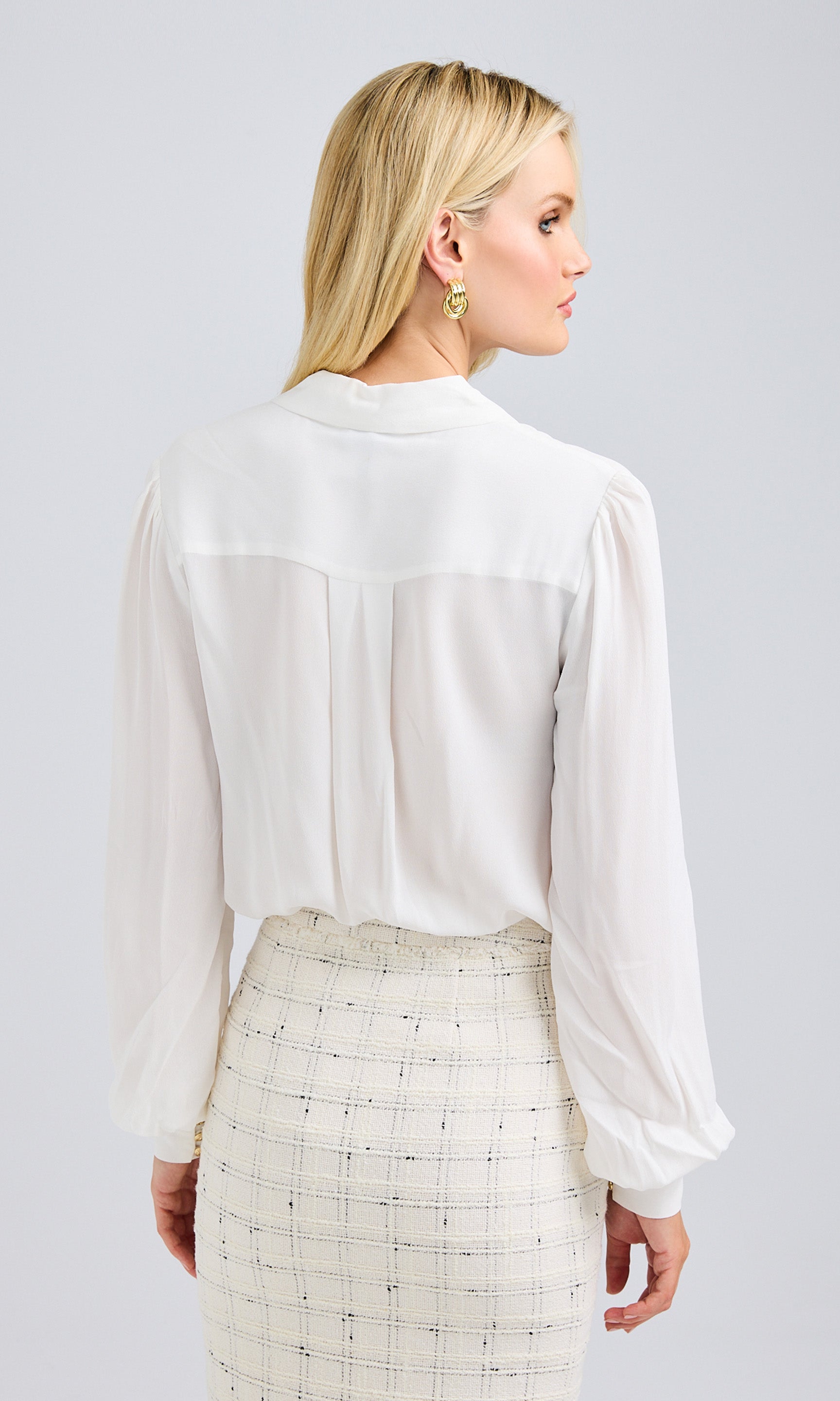 Rear view of a model wearing a white blouse with long sleeves and a tailored back design paired with a white tweed skirt.