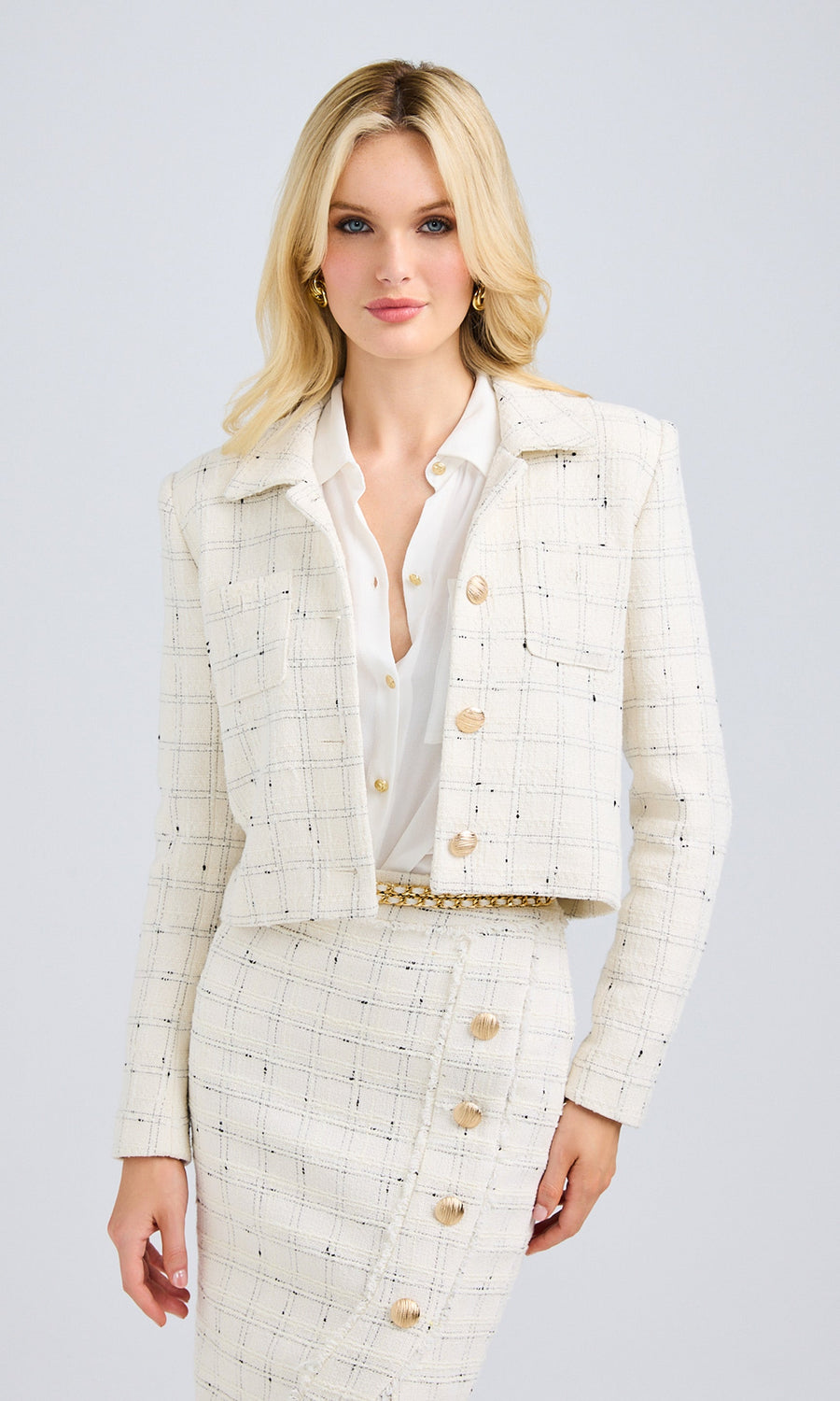 Model wearing a white tweed cropped jacket with gold laurel wreath shield buttons, paired with a matching tweed skirt.