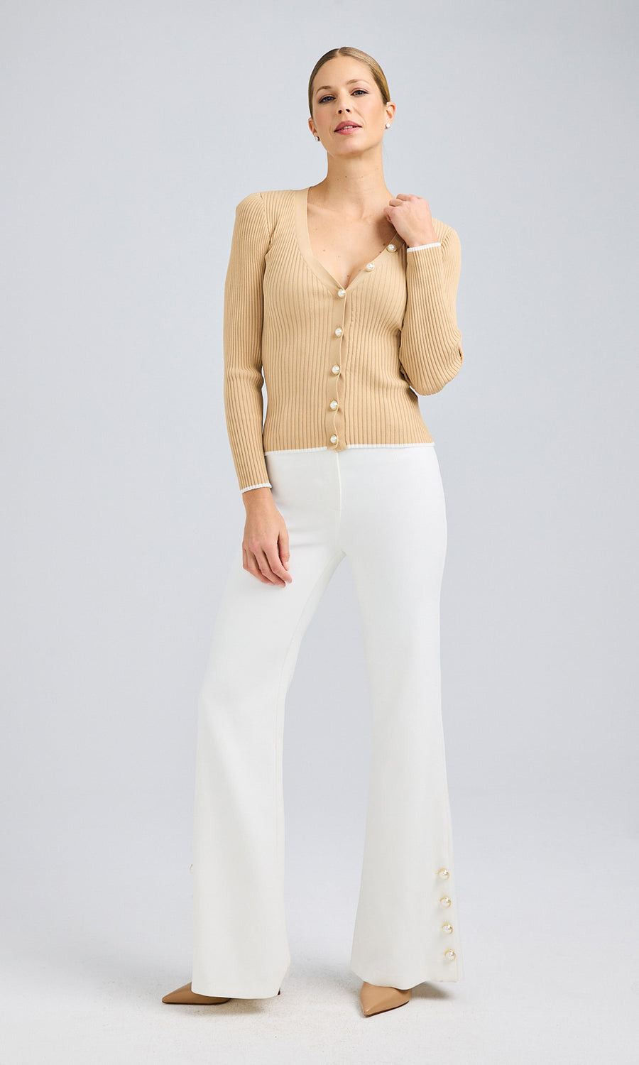 Full-body front view of a model wearing a beige ribbed cardigan with pearl buttons and white crepe pants with gold and pearl button embellishments along the hem.