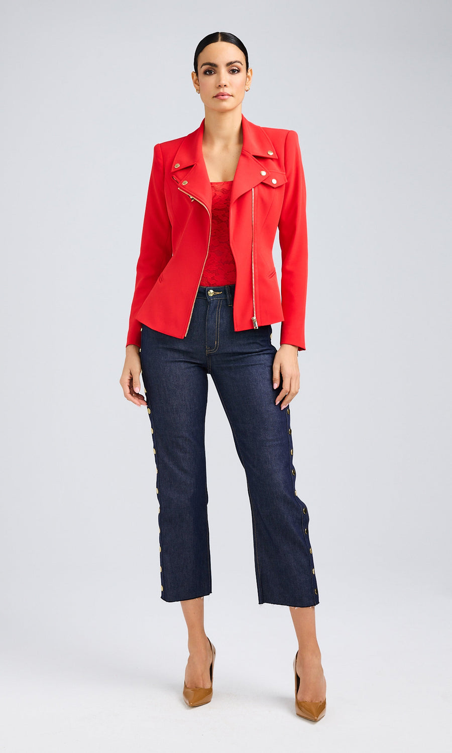 A brunette woman in a red moto jacket with gold details and jeans stands confidently.