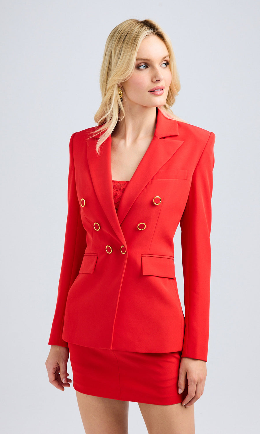 A woman in a red blazer with gold buttons and high heels stands confidently, showcasing her stylish outfit.