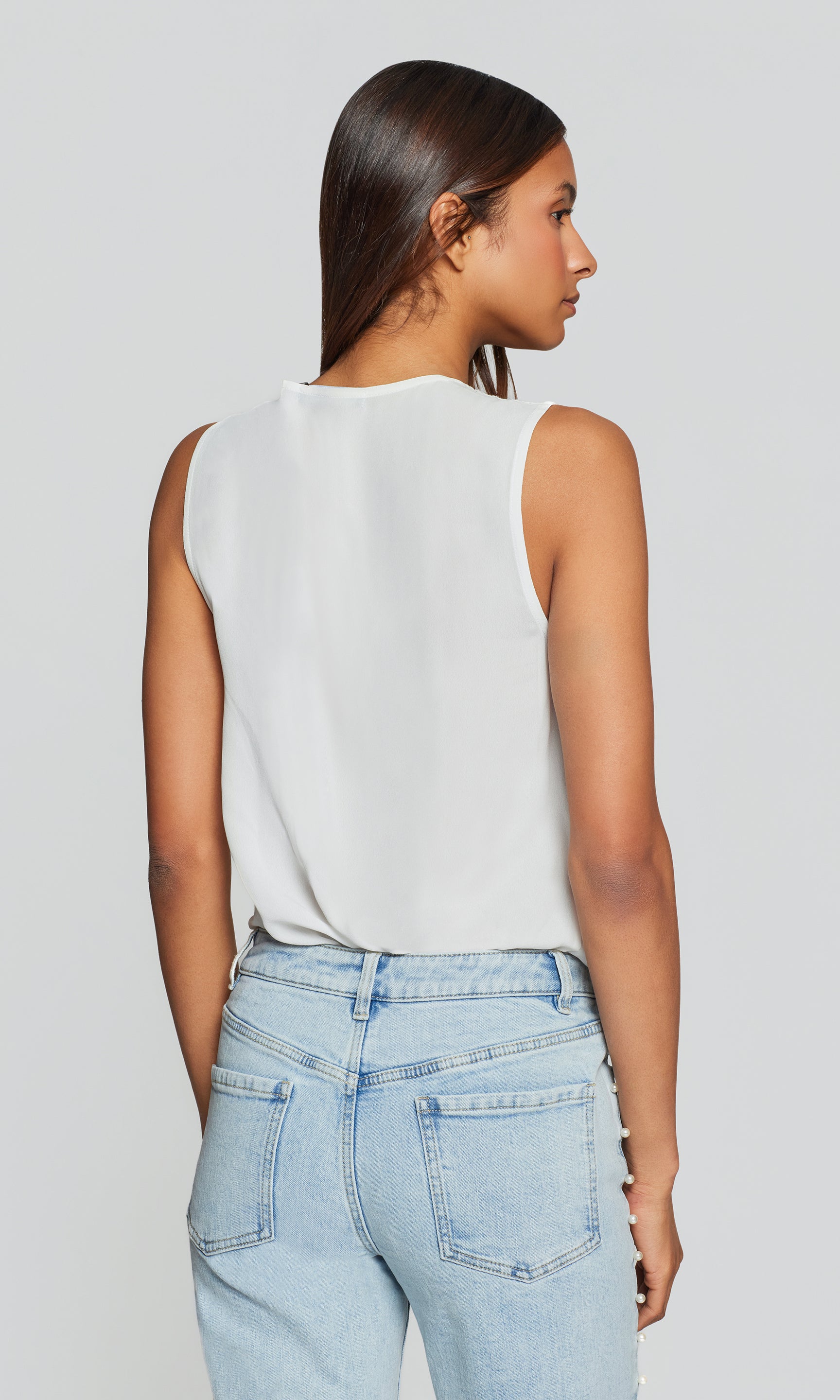Back view of woman wearing a white sleeveless top and light blue jeans against a plain background
