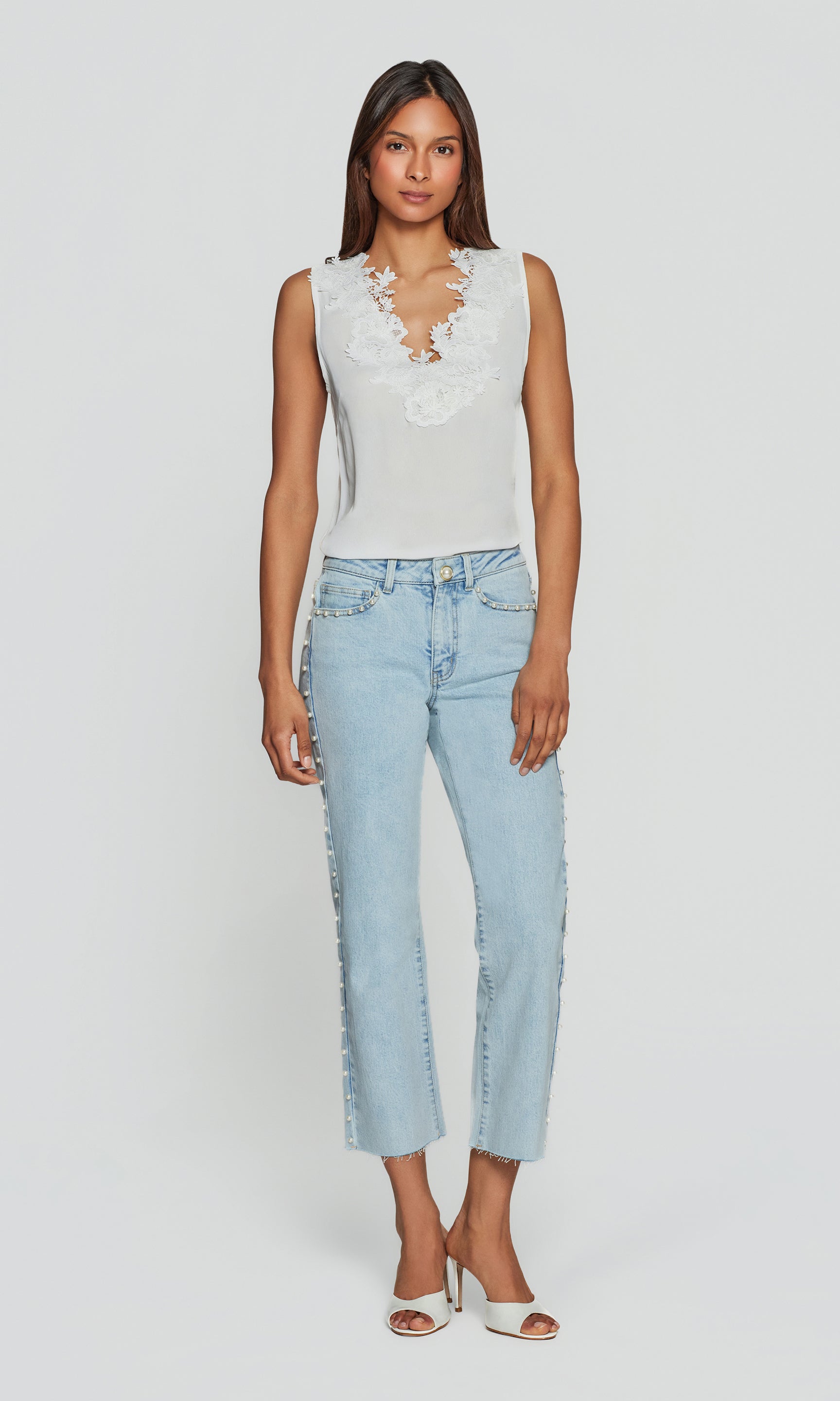 Woman wearing a white sleeveless top with lace details and light blue jeans on a white background